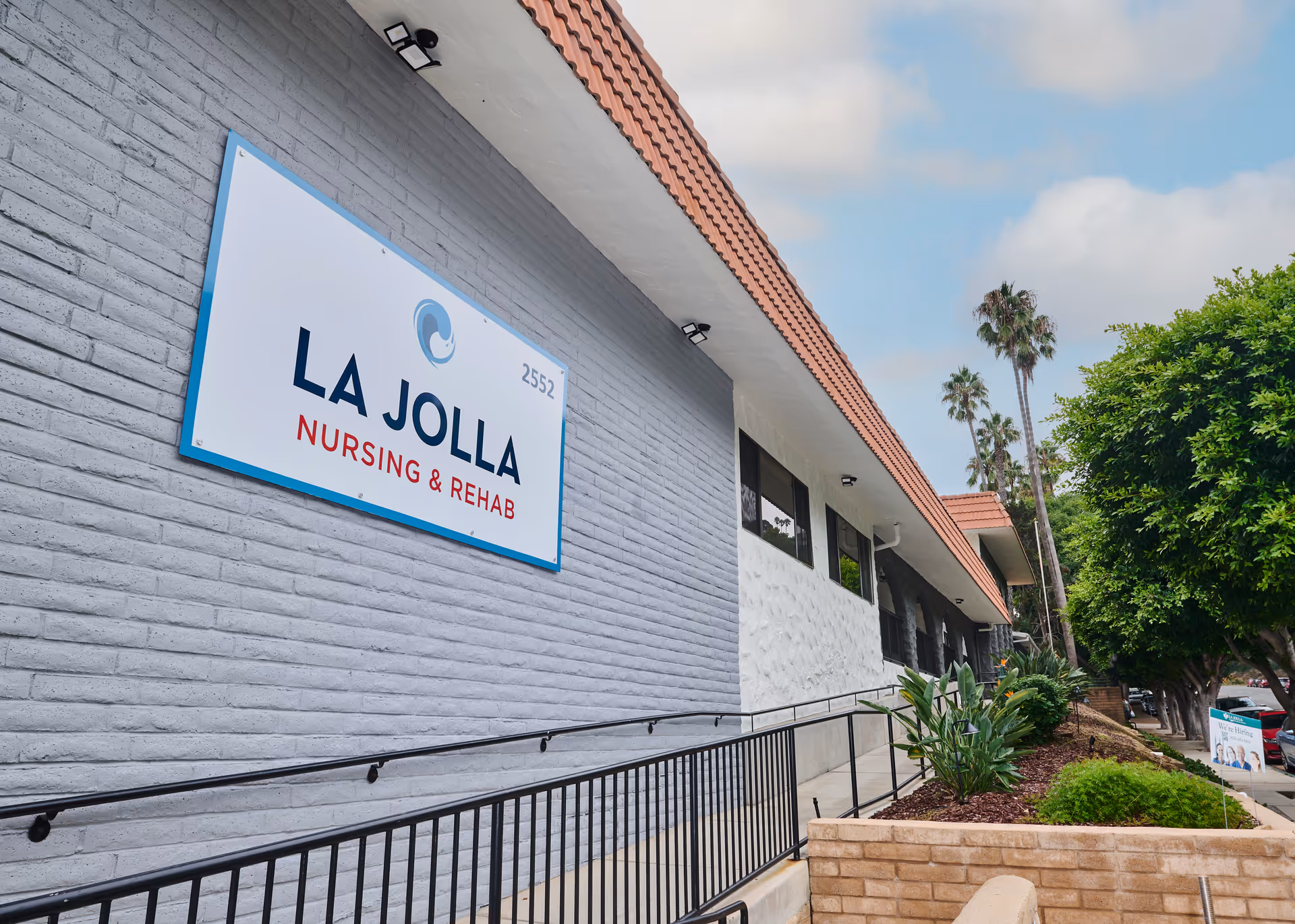 Exterior front of the La Jolla Nursing & Rehab building showing a large sign, wheelchair ramp, landscaping and palm trees.