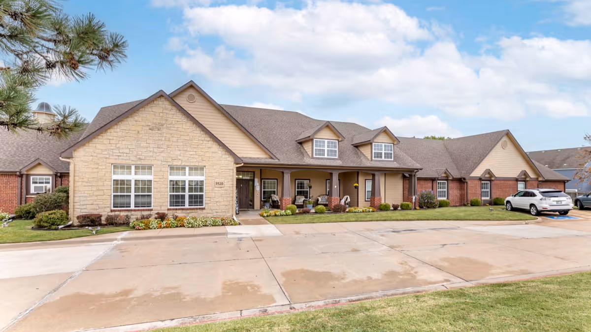 Exterior view of a single-story senior living facility building with a combination of stone and brick facade, multiple windows, a covered porch with seating, a driveway, and parked cars under a partly cloudy sky.