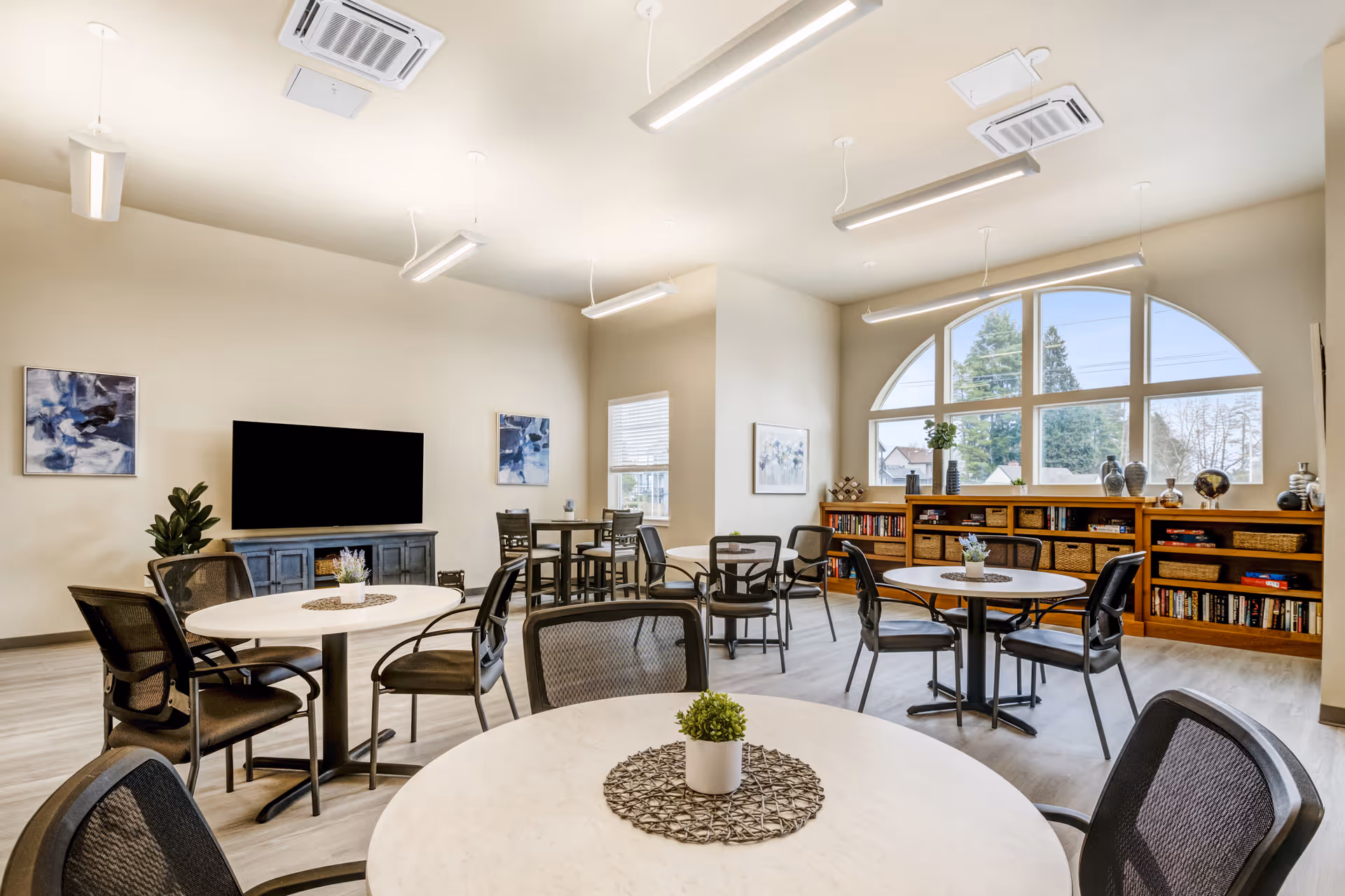 A bright and spacious common area with multiple round tables and black mesh chairs arranged around them. The room features large arched windows letting in natural light, a wooden bookshelf filled with books and decorative items, a flat-screen TV mounted on a blue cabinet, and modern ceiling lights. The walls are decorated with abstract paintings, and small potted plants are placed on the tables.