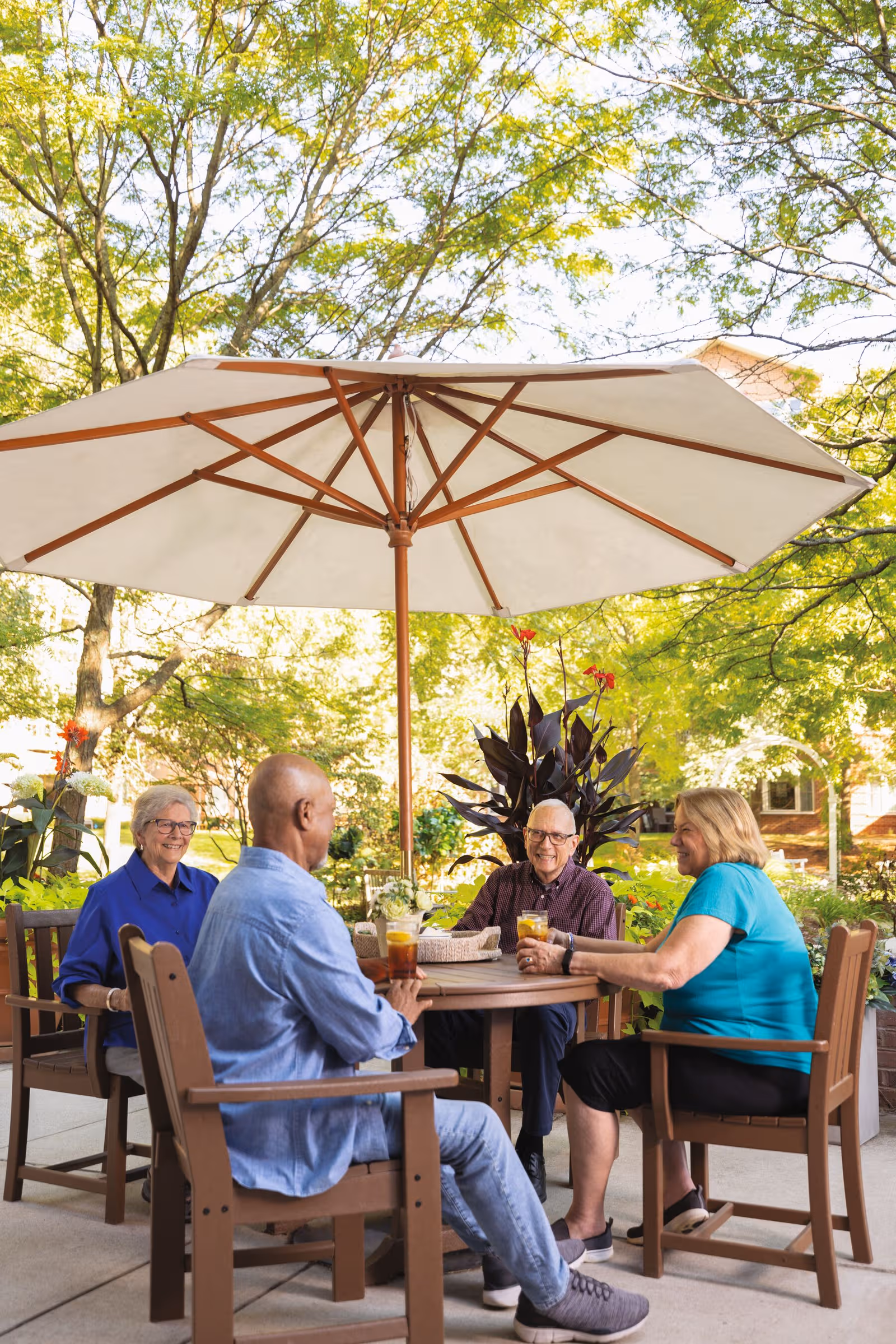 Four seniors sit around a round outdoor table under a large patio umbrella in a courtyard.