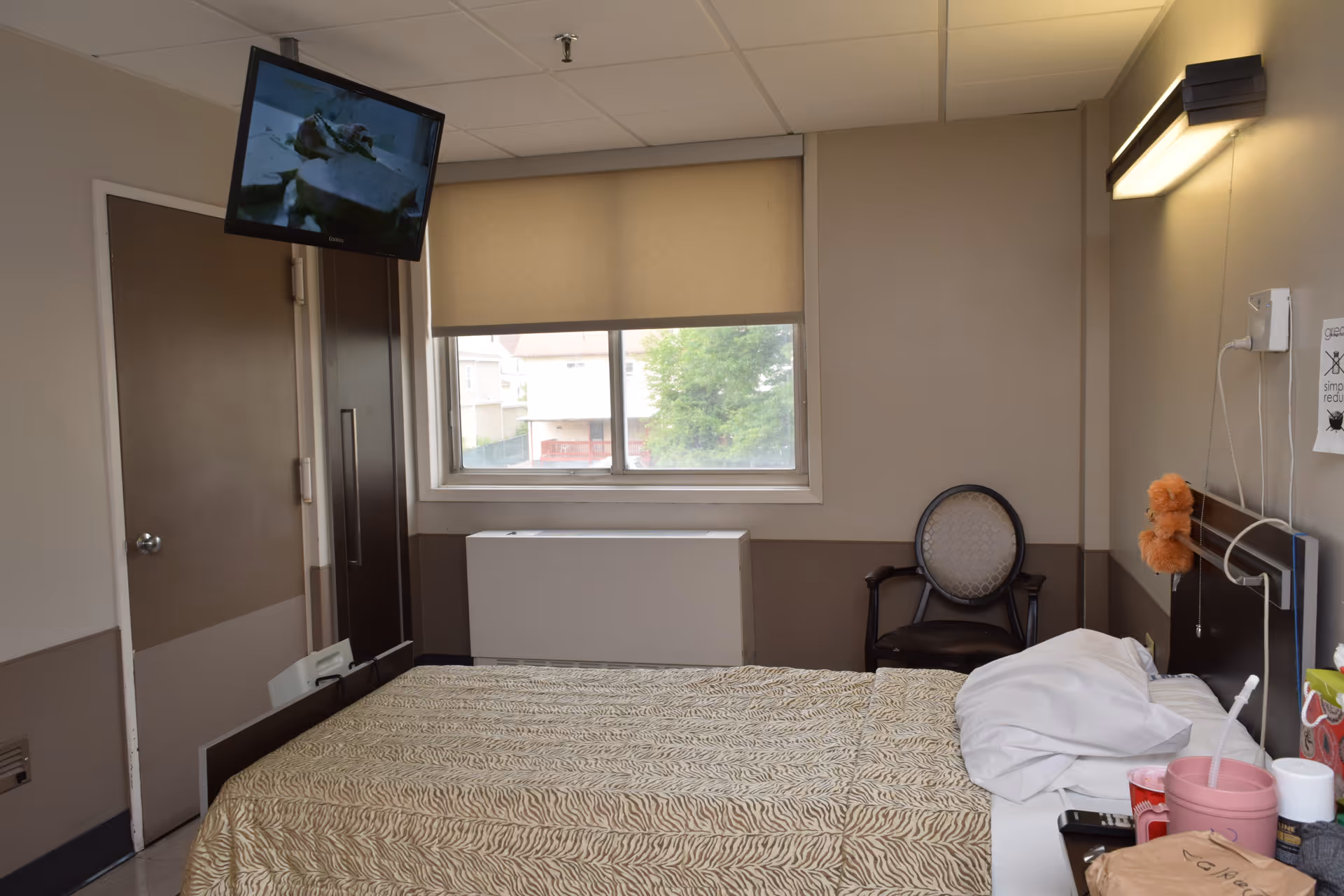 Hospital-style patient room with a bed, window, wall-mounted TV, and chair.
