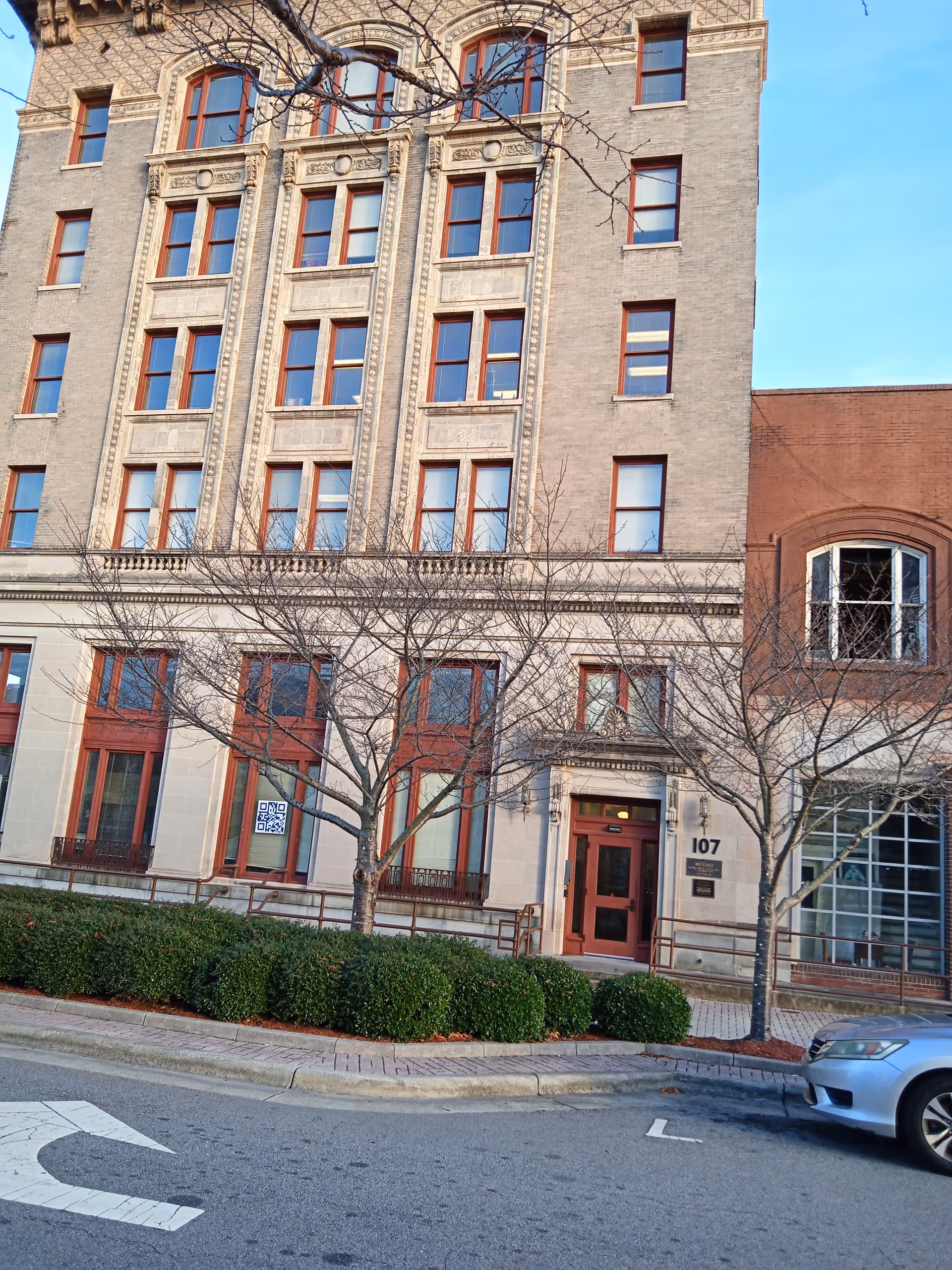 Brick, multi-story building with arched windows and a front entrance, trees along the sidewalk.