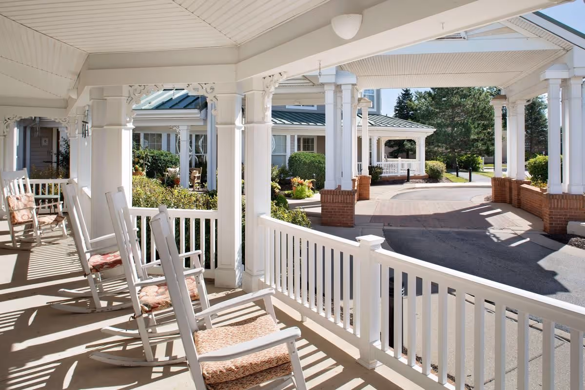 Covered porch with white rocking chairs and railings overlooking a porte-cochère entrance, driveway, and landscaped grounds.