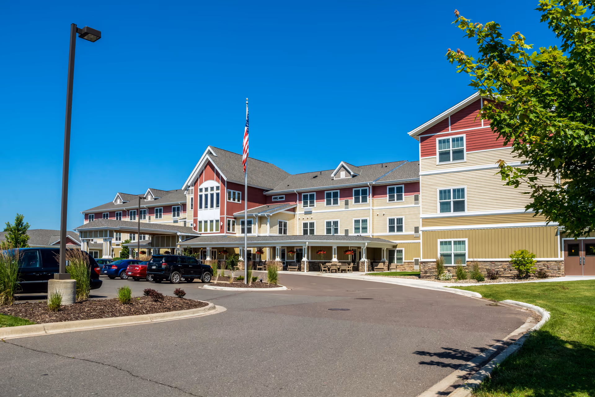 Exterior view of a senior living facility building with a driveway and parked cars in front. The building has multiple stories with beige, red, and yellow siding, several windows, and a covered entrance area. An American flag is displayed on a flagpole near the entrance. The sky is clear and blue, and there is greenery including trees and shrubs around the building.
