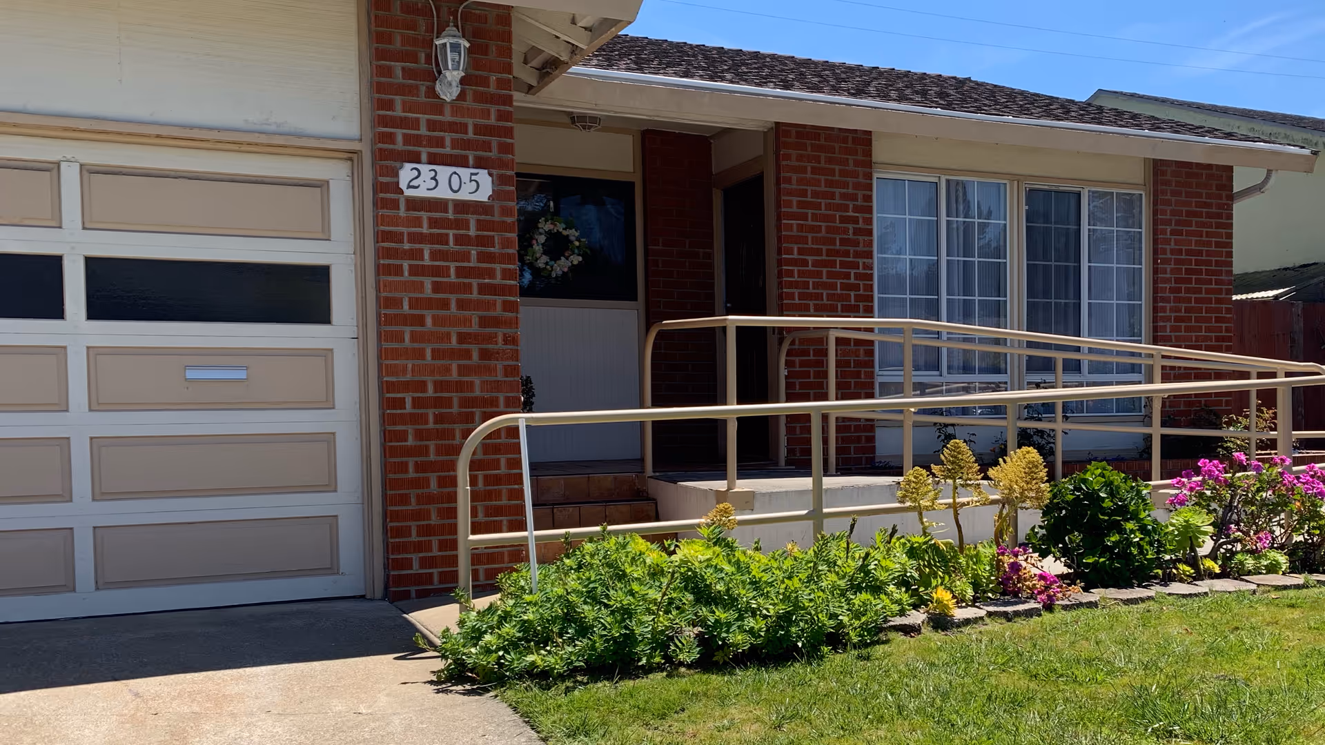 Front entrance of Heirloom Rose Garden senior living facility, brick building with ramp and flowering garden.