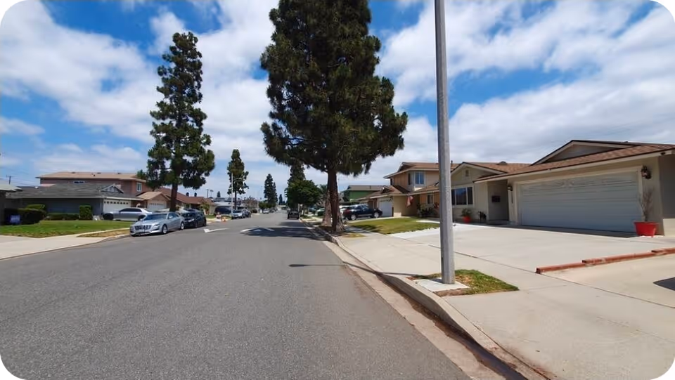 Residential street scene with single-story homes, trees, and parked cars.
