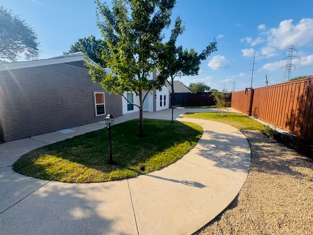 Curved sidewalk winds through a grassy courtyard with trees beside a single-story brick building.