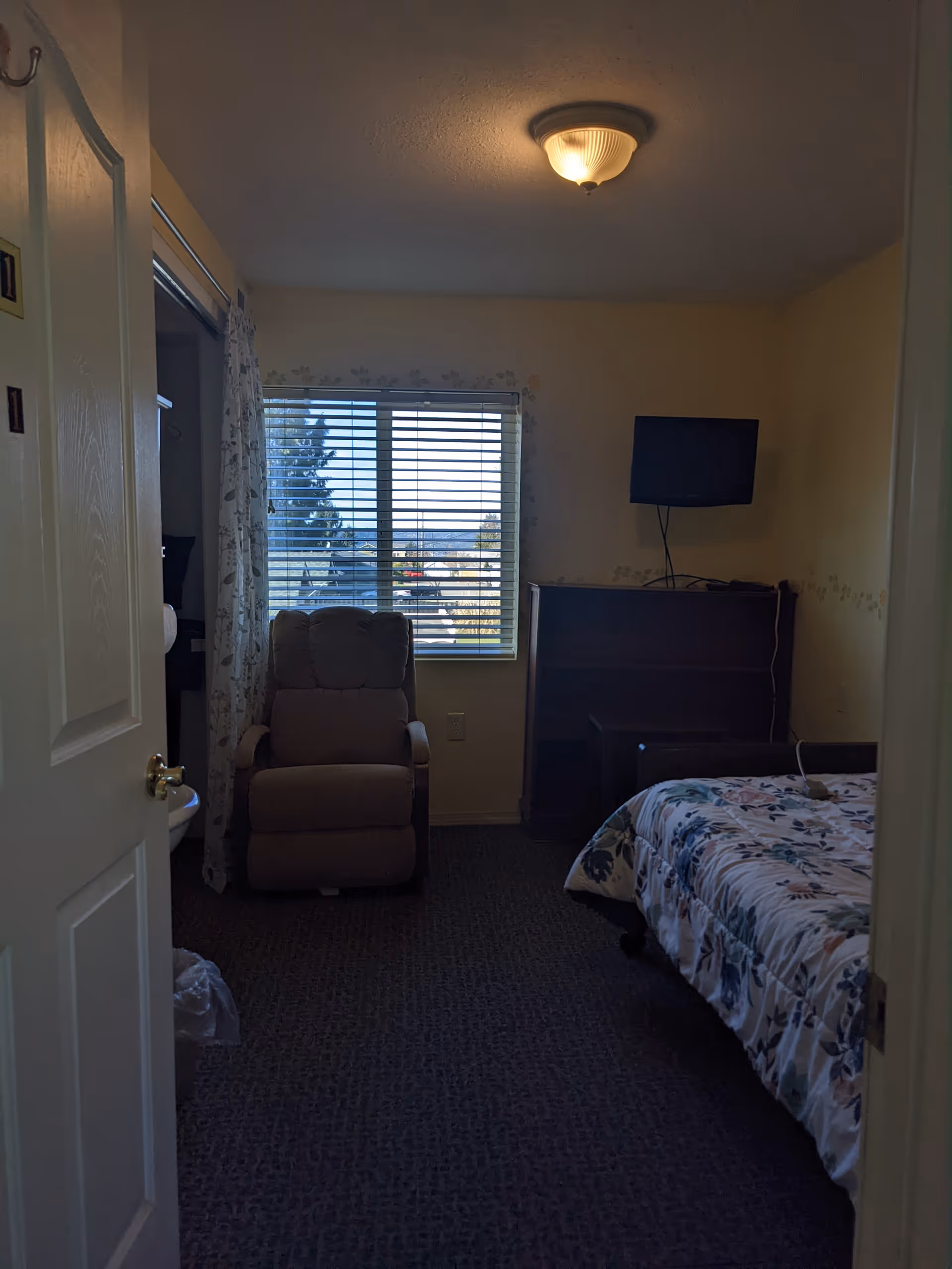 A furnished bedroom with a bed, recliner chair, dresser, and window blinds.
