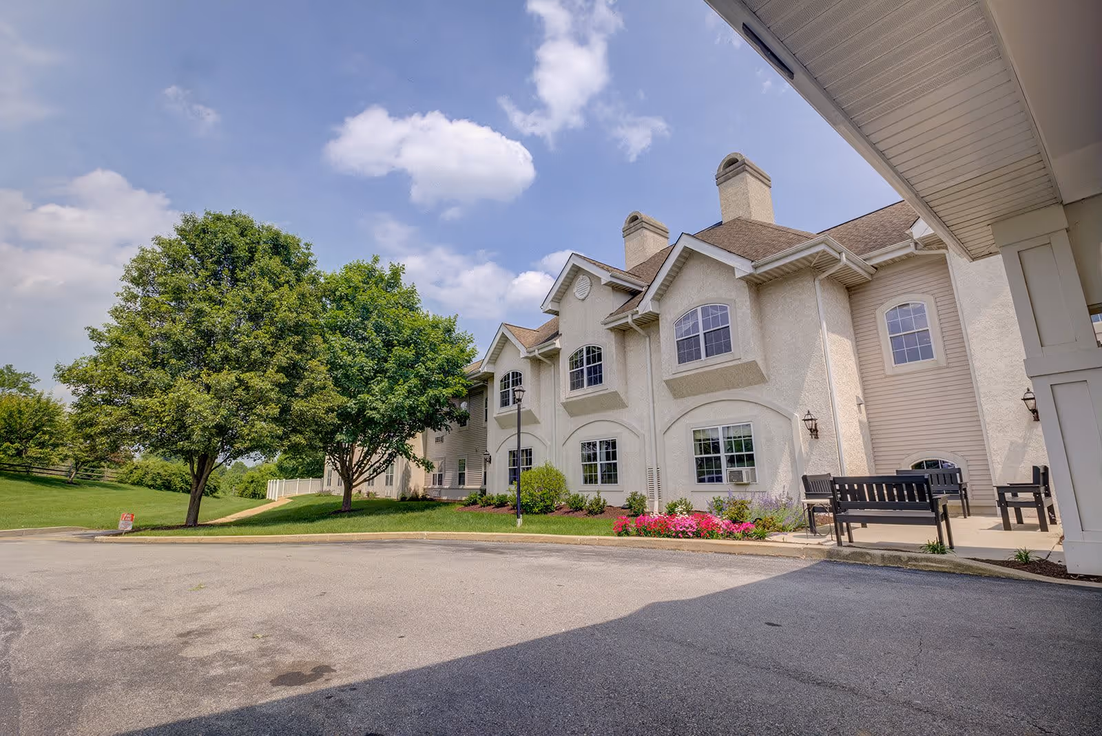 Exterior view of a two-story senior living facility building with beige walls and multiple windows. There are benches on a paved area near the entrance, a large green tree, and a well-maintained lawn with flowers under a partly cloudy sky.