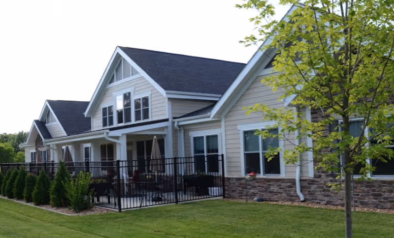 Exterior view of a light-colored multi-gabled building with fenced patios, a manicured lawn, and small trees.