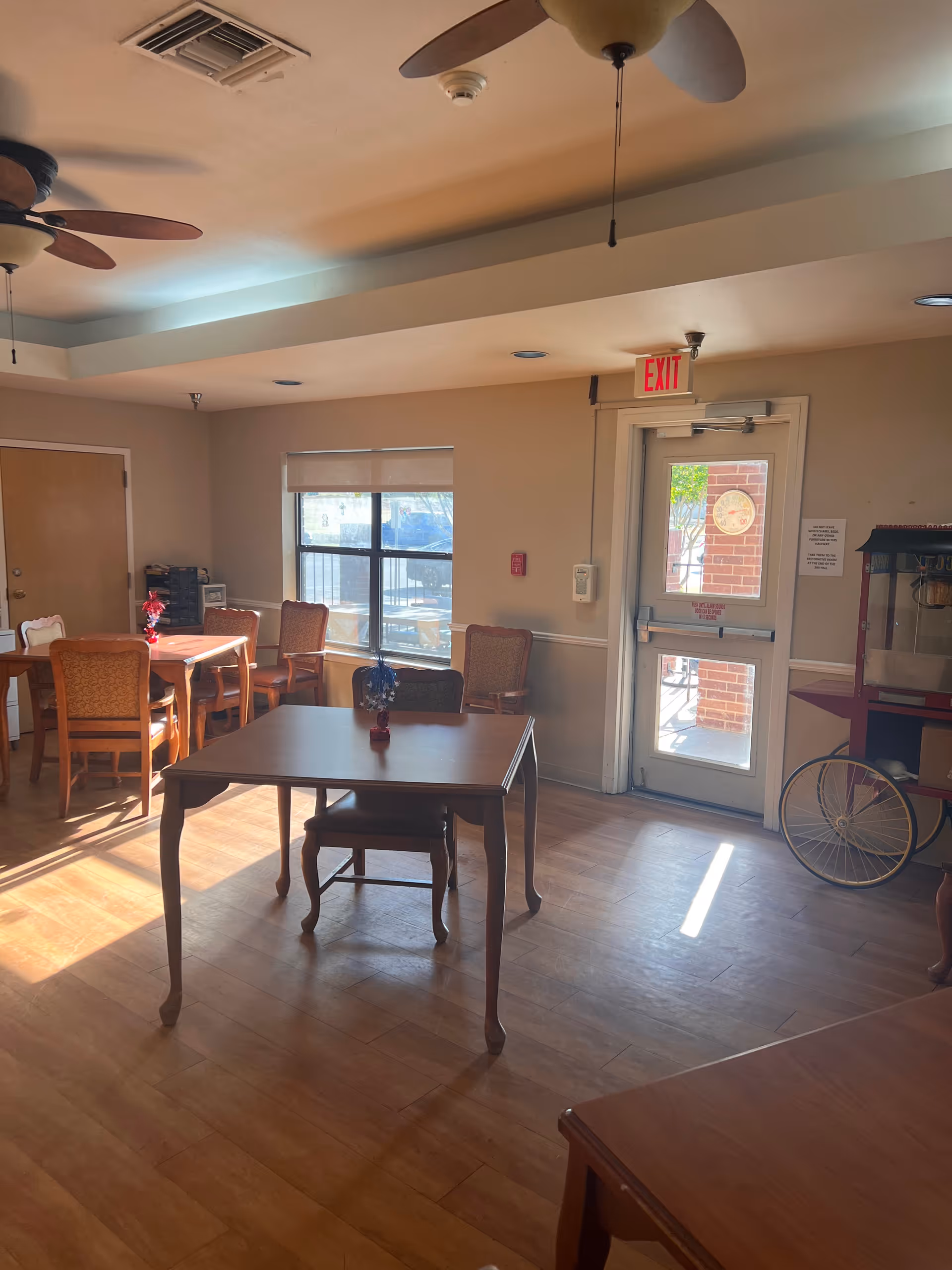 A well-lit room with wooden floors and several tables and chairs arranged around the space. There is a window letting in natural light, an exit door with a red exit sign above it, and a popcorn machine on the right side. Ceiling fans are mounted on the ceiling.