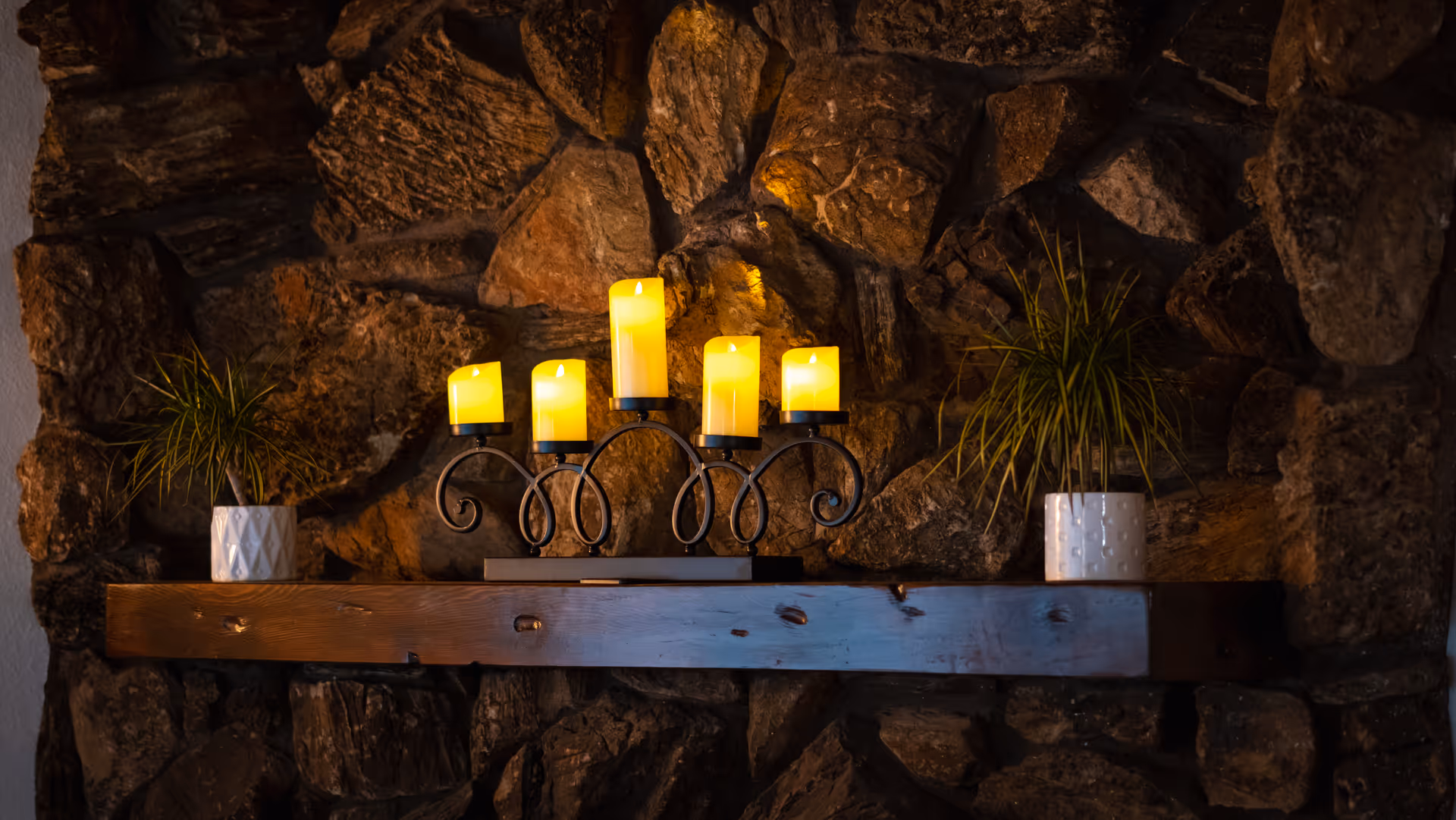 A rustic stone wall with a wooden shelf mounted on it. On the shelf, there is a black metal candle holder with five lit yellow candles and two small potted plants in white pots placed on either side of the candle holder.