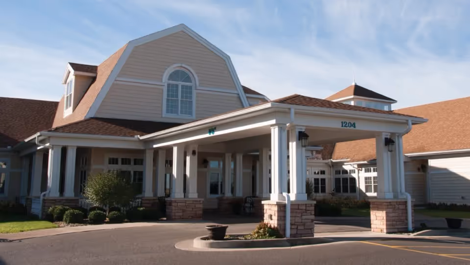 Exterior view of a senior living facility building with a covered entrance supported by white columns and stone bases. The building has beige siding, multiple windows, and a brown shingled roof under a partly cloudy blue sky.