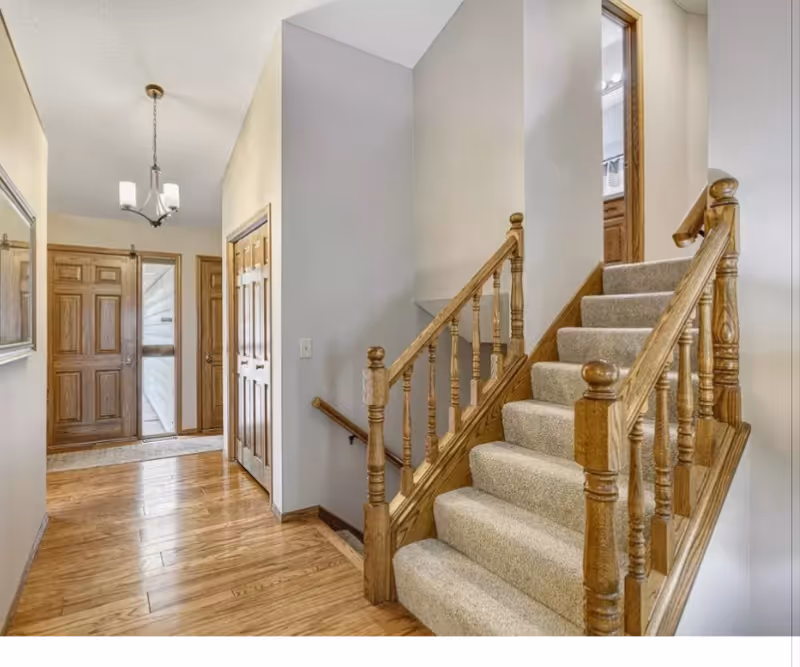 Bright hallway with a wooden staircase, carpeted steps, and front entry doors.