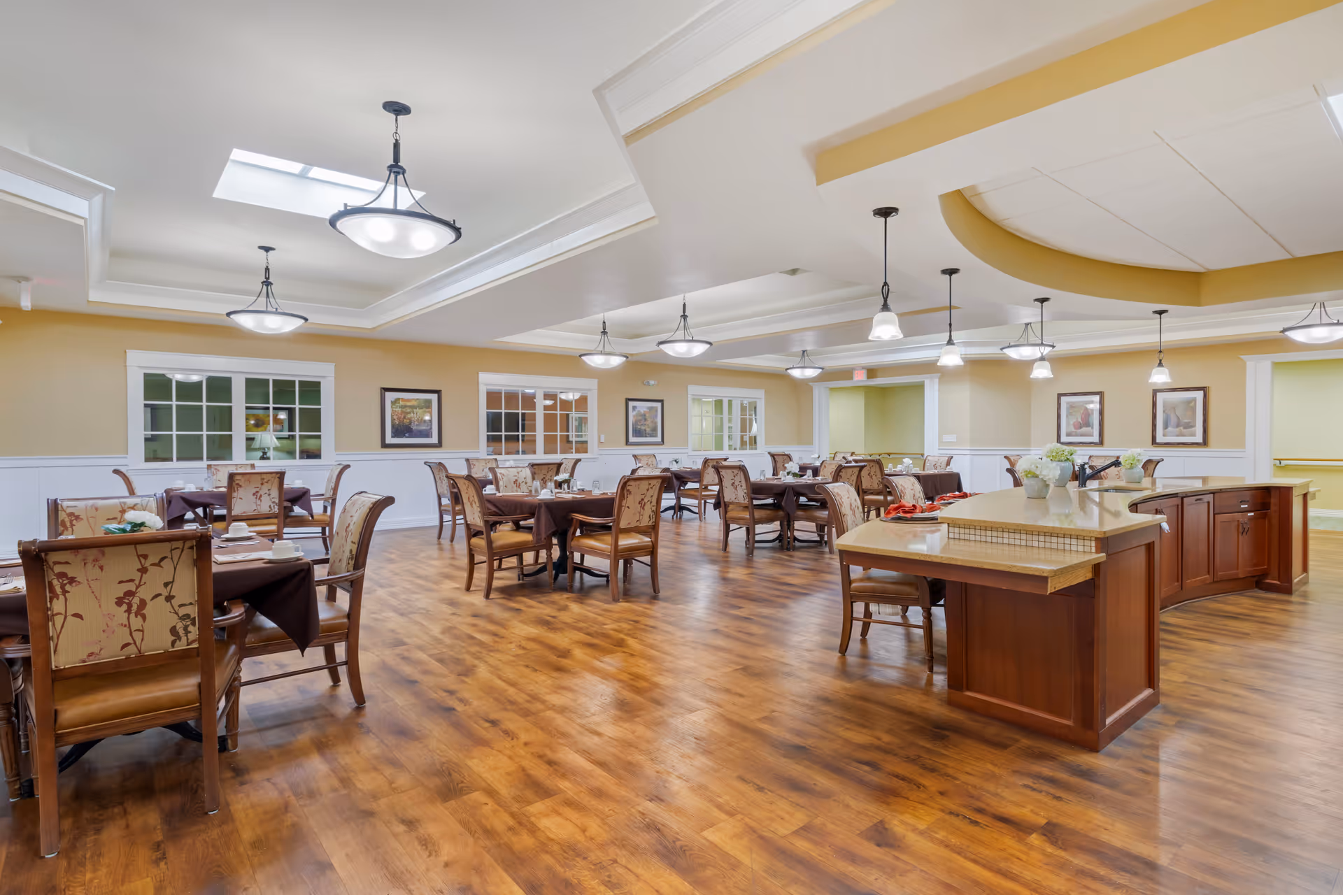 A spacious dining room with wooden floors, multiple tables covered with dark tablecloths, and cushioned chairs with floral upholstery. The room features a large island counter with a beige countertop and wooden cabinetry. The ceiling has recessed lighting and hanging pendant lights, with framed artwork on the walls and windows looking into adjacent rooms.