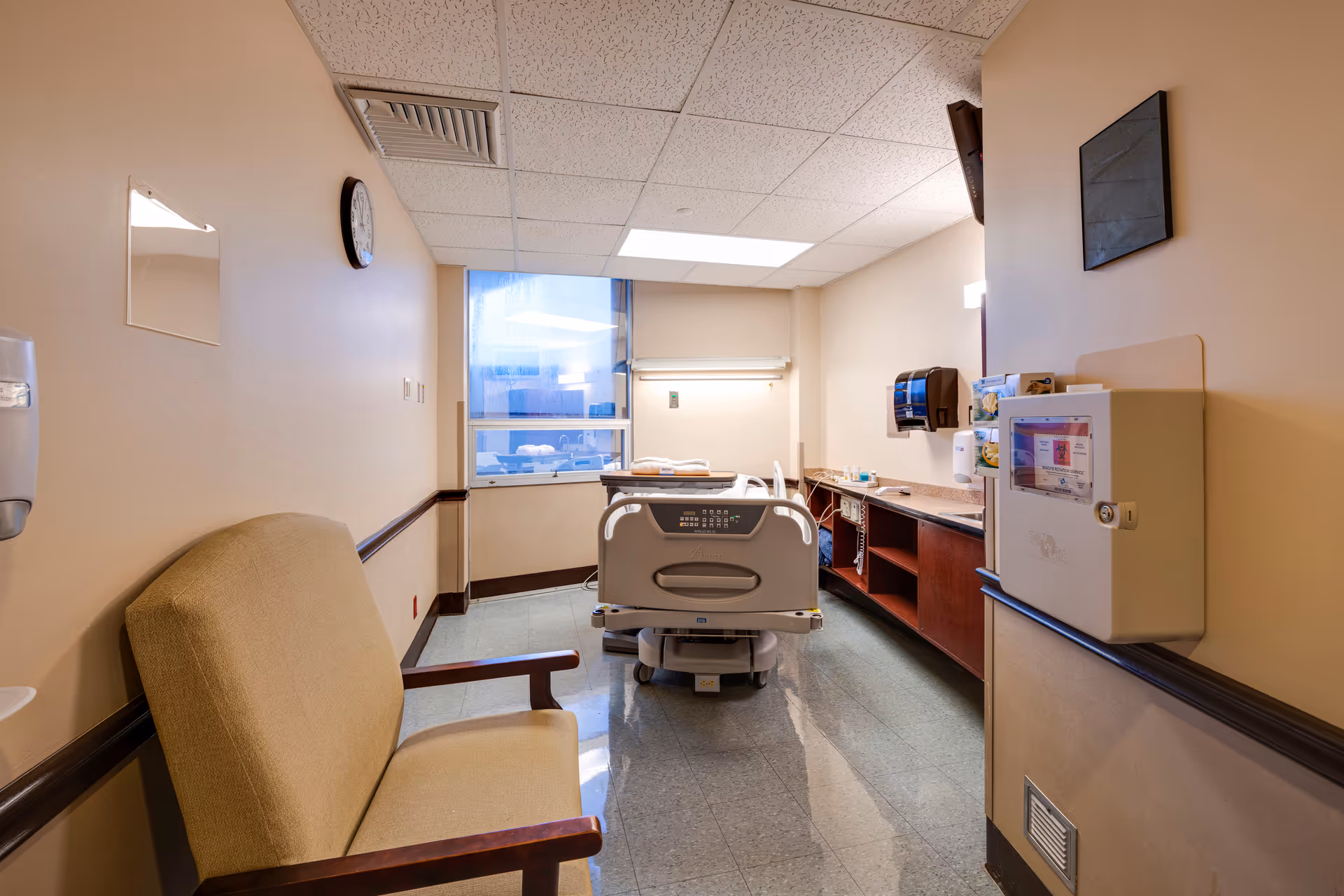 Hospital bed in a clinical interior with chair, cabinets, and window.