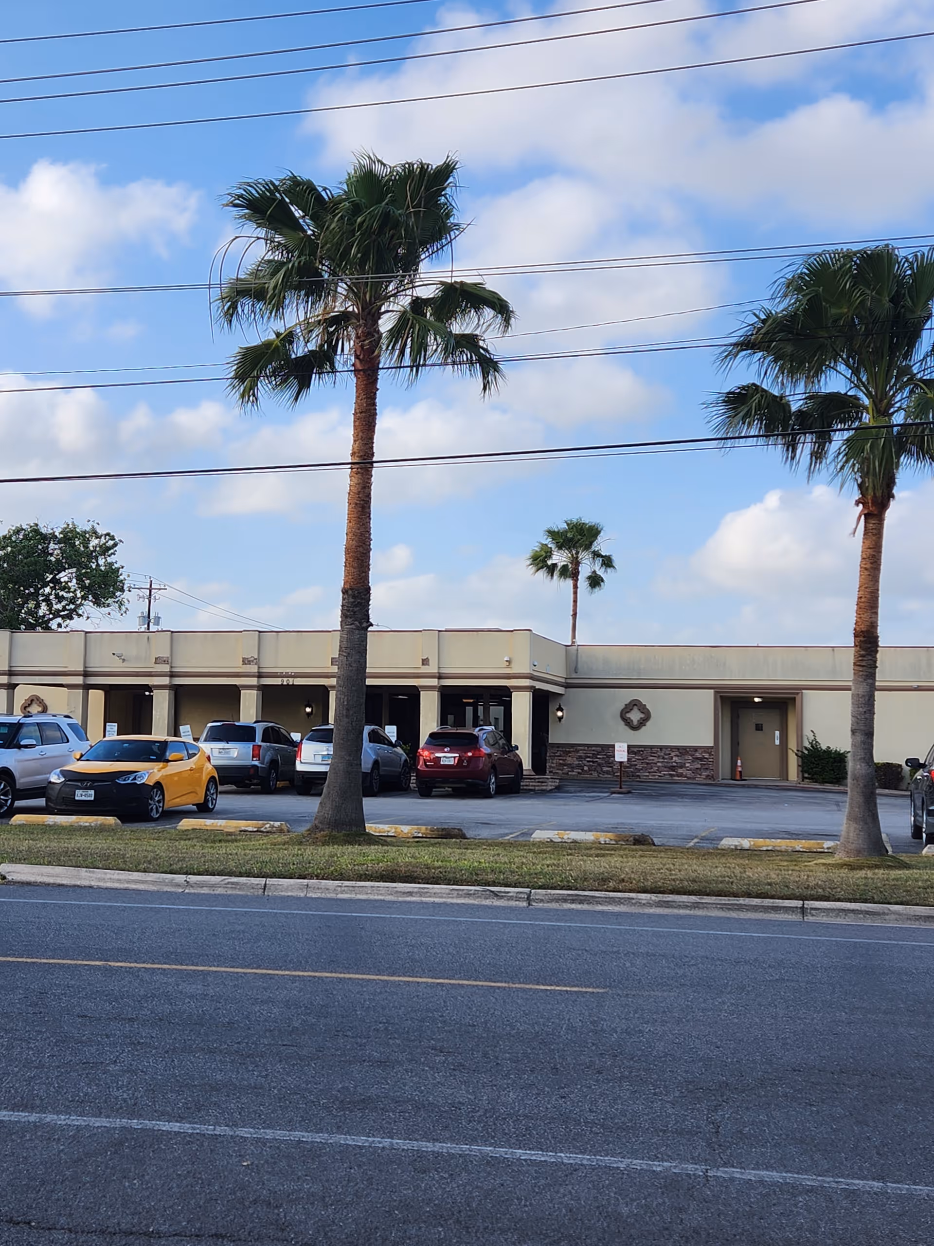 Exterior view of a single-story senior living facility with palm trees and parked cars.