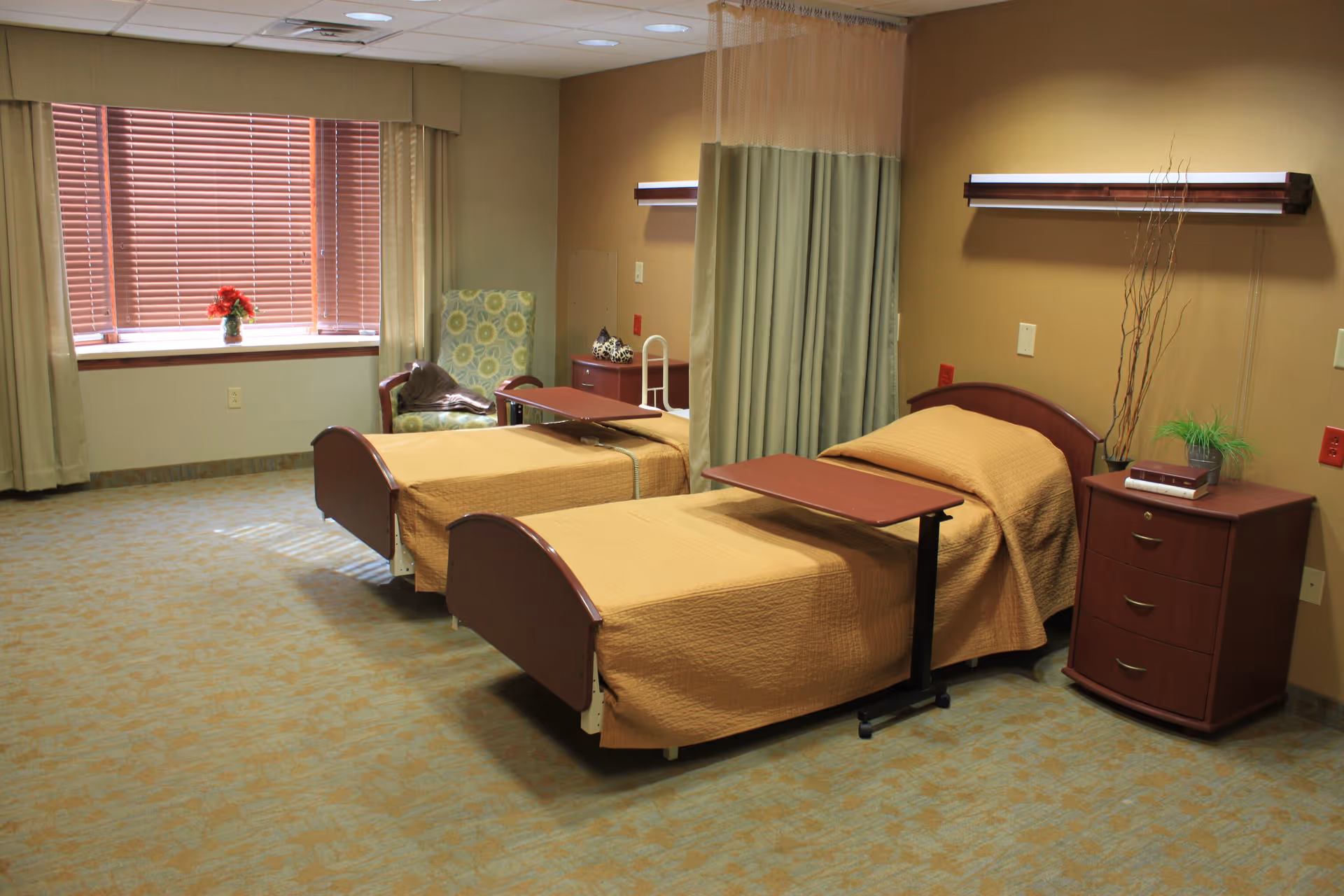 Two hospital-style beds with tan bedspreads, overbed tables, a curtain divider, bedside furniture, and a window with blinds in a senior living facility room.