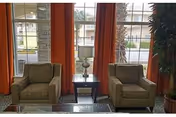 A seating area with two beige armchairs, a small table, lamp, and red curtains.