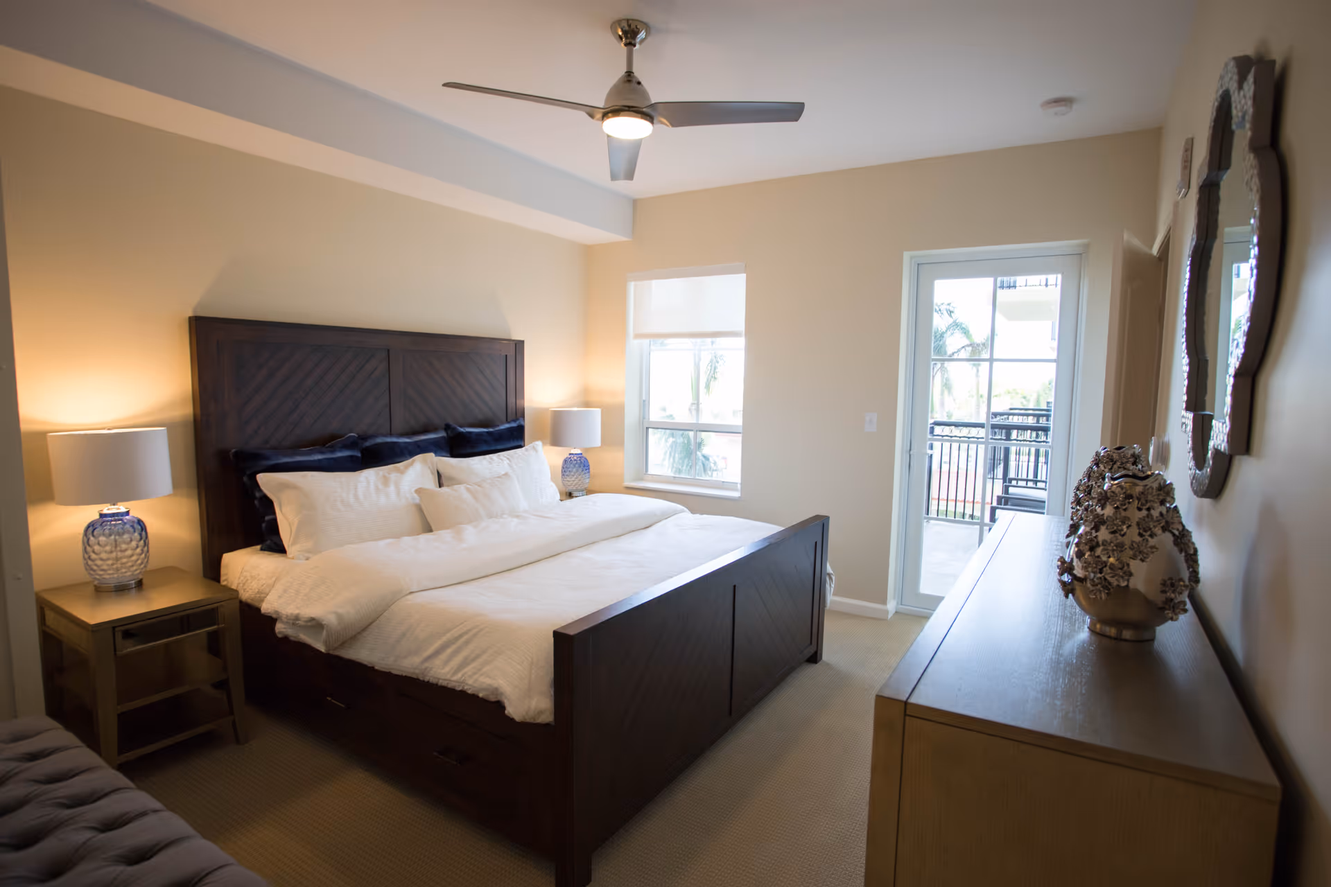Bright bedroom with a large wooden bed, bedside tables and lamps, a dresser, and a door leading to a balcony.