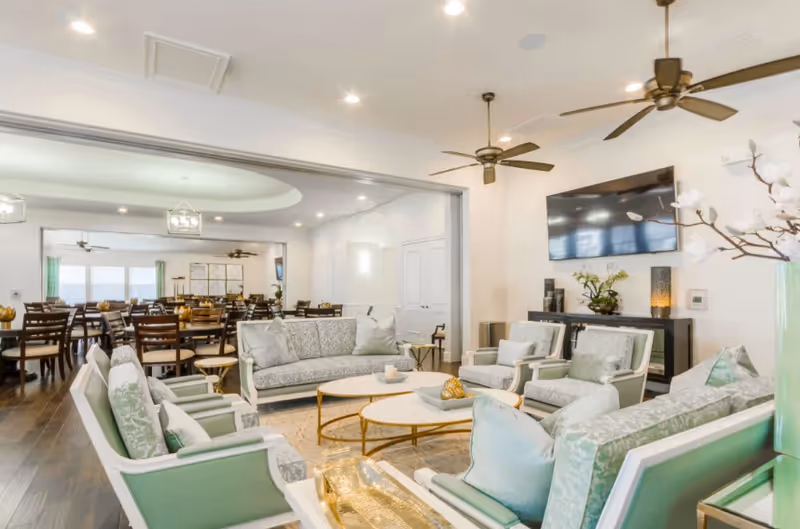 Bright senior living lounge with mint-green seating, a wall-mounted TV, and open dining area.