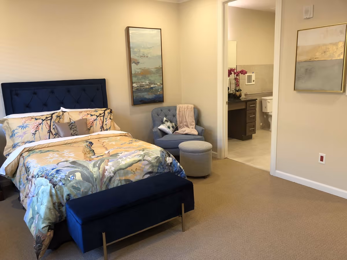 Windhaven Senior Living bedroom with blue upholstered headboard, seating area, and adjacent bathroom.