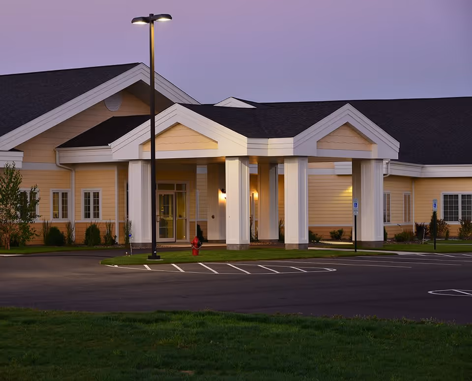 Exterior view of a senior living facility building at dusk with a covered entrance supported by white columns, a lit streetlamp, and a parking lot with marked spaces and handicap signs.