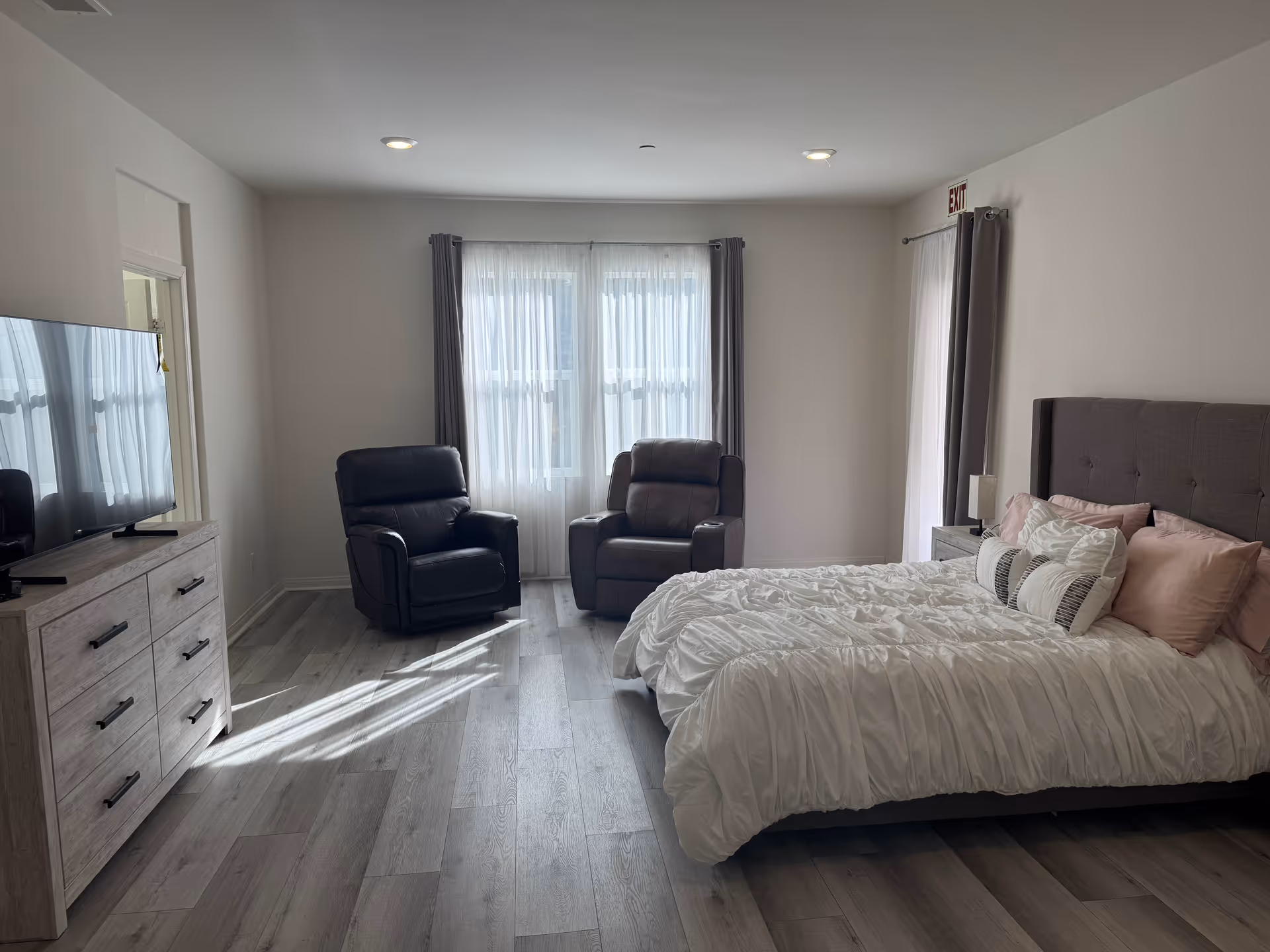 A bright bedroom with a large bed featuring white bedding and multiple pillows, two dark recliner chairs near a window with sheer white curtains and gray drapes, a wooden dresser with a flat-screen TV on top, and light wood flooring.