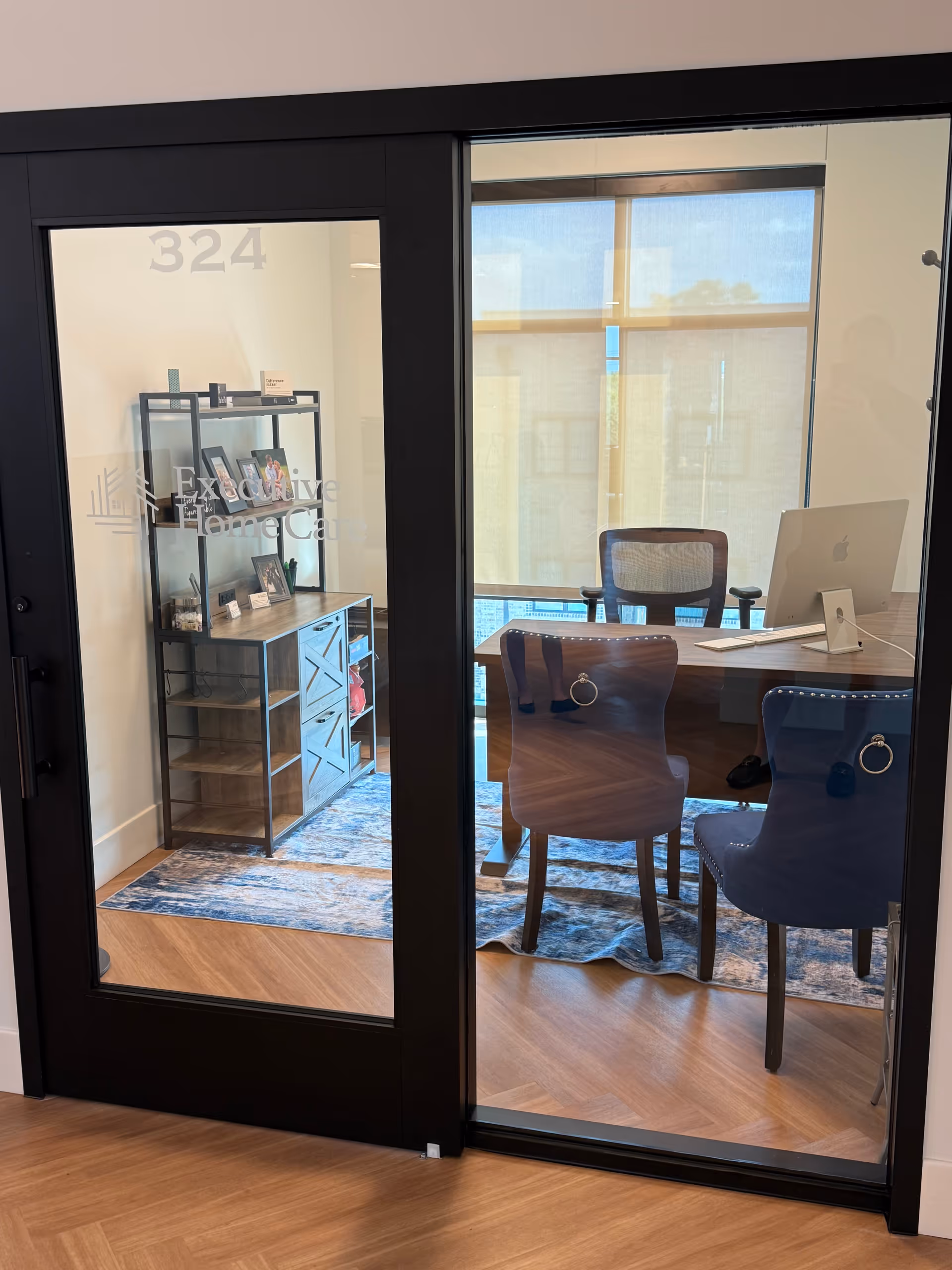 Glass-walled office with desk, chairs, and shelving inside a facility.