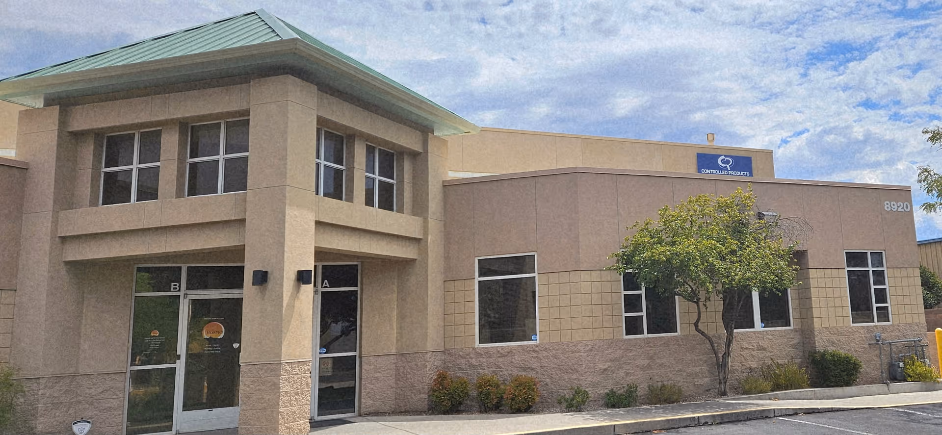 Exterior view of a beige facility building with glass doors and a small tree.