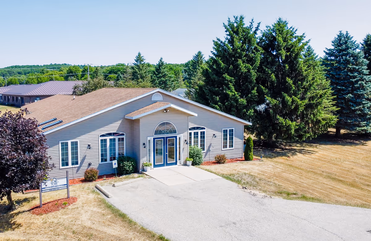 Exterior view of a single-story senior living facility building with beige siding and a brown roof, surrounded by trees and a grassy area under a clear blue sky.