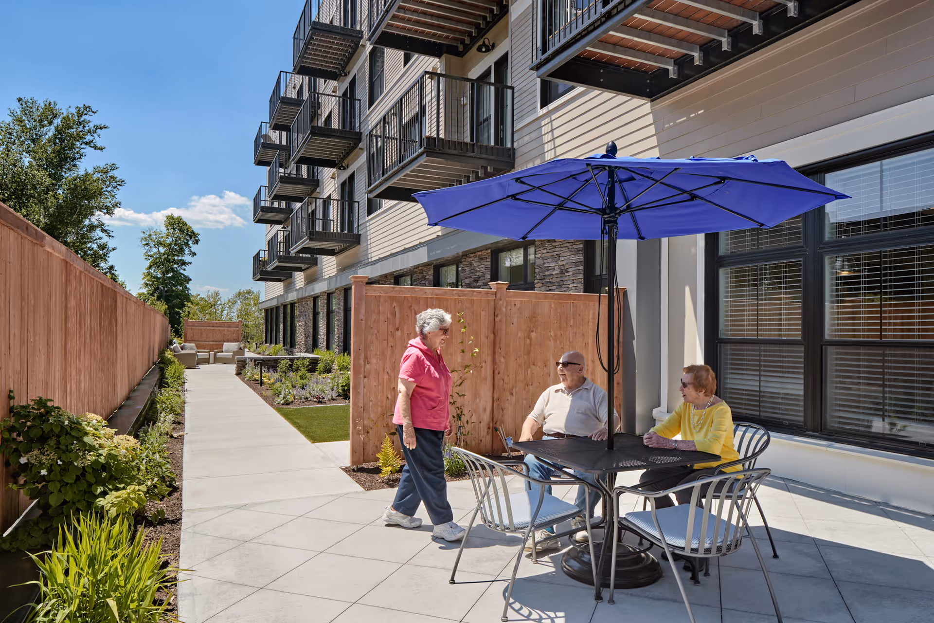 Three senior residents sit and chat at a patio table under a blue umbrella next to a multi-story building with balconies.