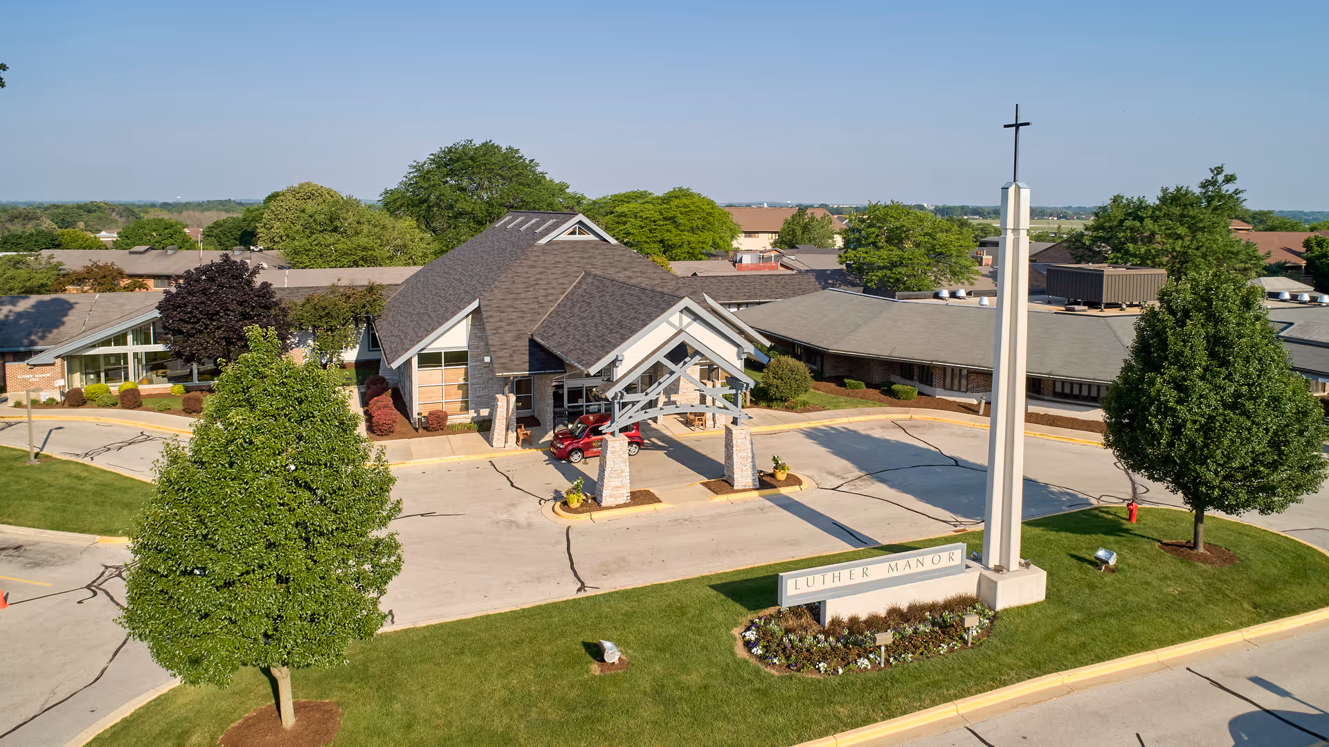 Aerial view of Luther Manor facility showing a large building with a peaked roof entrance, surrounded by trees and landscaped greenery. A tall white cross monument and a sign with the name Luther Manor are visible in the front lawn area.
