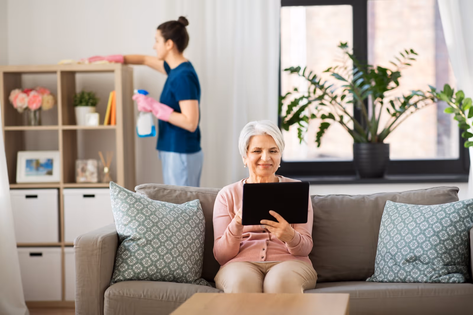 An elderly woman lounges on a sofa using a tablet while a caregiver cleans shelves.