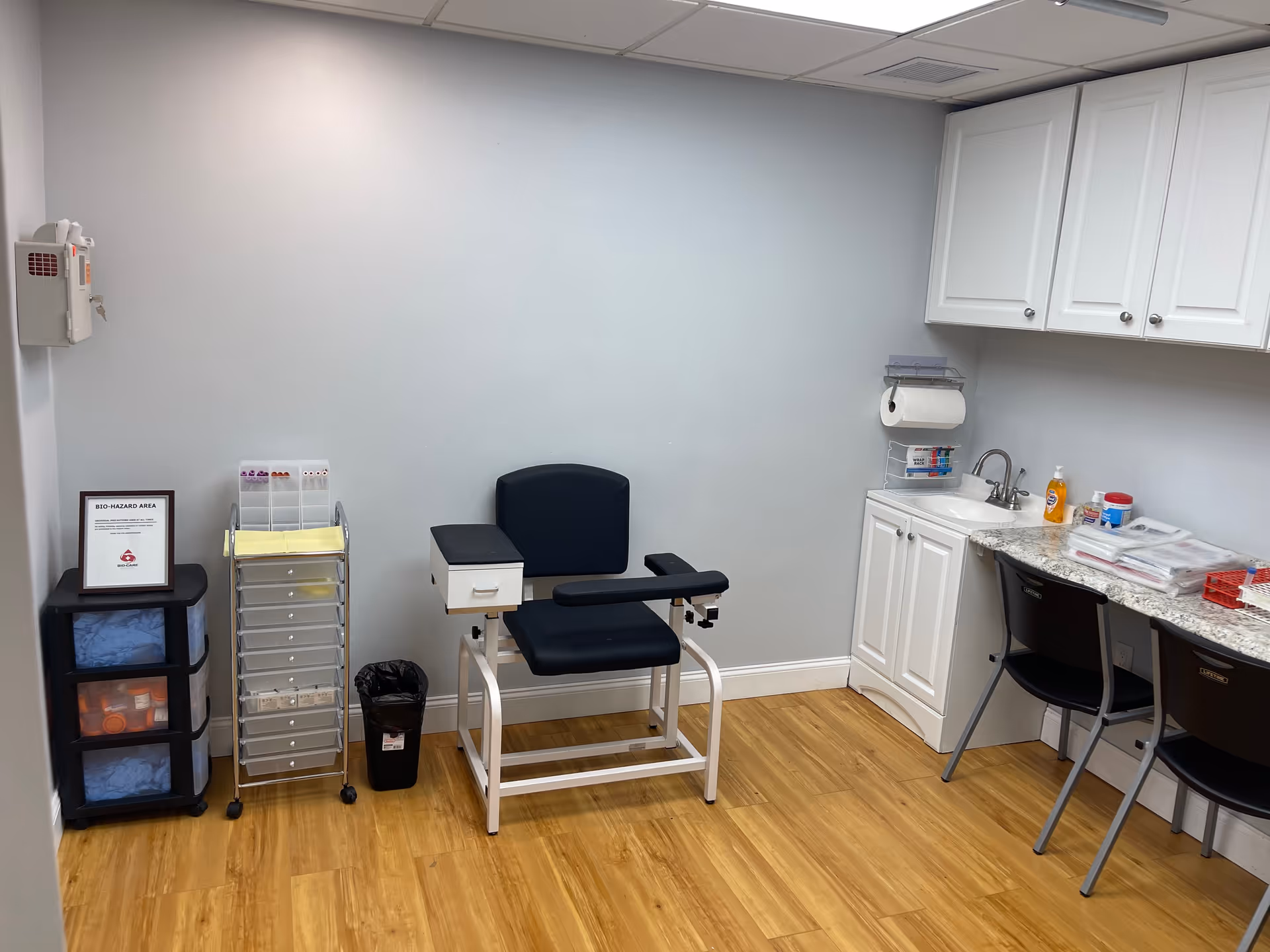 A small clinical room with a blue examination chair, sink, white cabinets, and medical supplies.