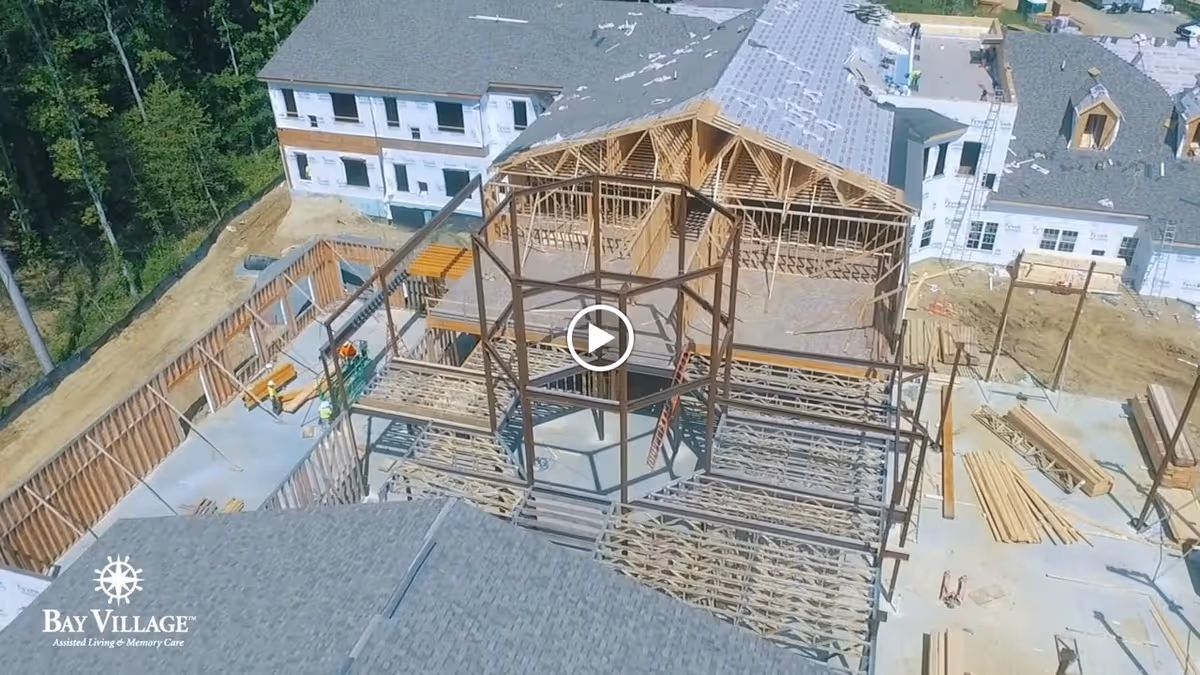 Aerial view of a construction site showing the framework and partially completed buildings of a senior living facility named Bay Village. The site includes wooden and steel structural elements, with some buildings having roofs and windows installed, surrounded by trees and dirt ground.