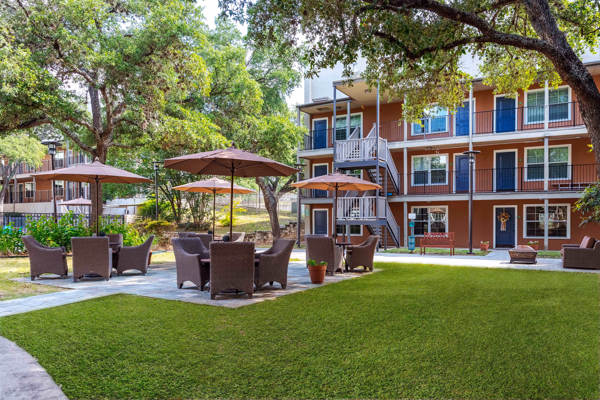 Outdoor courtyard with wicker seating and umbrellas in front of a three-story residential building surrounded by trees.