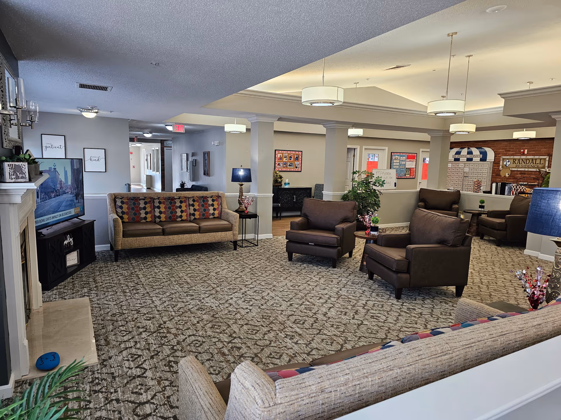 A spacious living room area in a senior living facility with patterned carpet, multiple armchairs and sofas with colorful cushions, a flat-screen TV on a stand, a fireplace, decorative lamps, and plants. The room has a high ceiling with recessed lighting and columns separating different sections of the space.