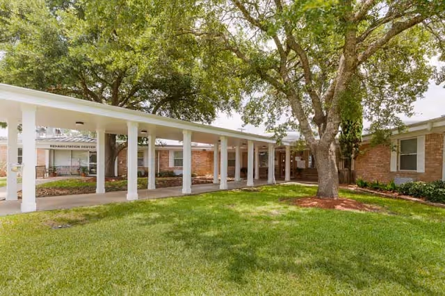 Single-story brick senior living facility entrance with a covered white-column walkway, green lawn, and large trees.