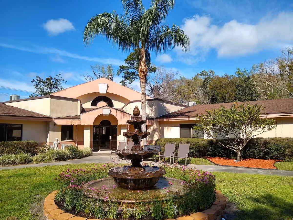 Exterior view of Diamond Assisted Living & Memory Care facility featuring a beige building with a brown roof, a central entrance with an archway, a tall palm tree, a multi-tiered water fountain surrounded by flowers, and outdoor seating chairs on a sunny day with a blue sky and scattered clouds.