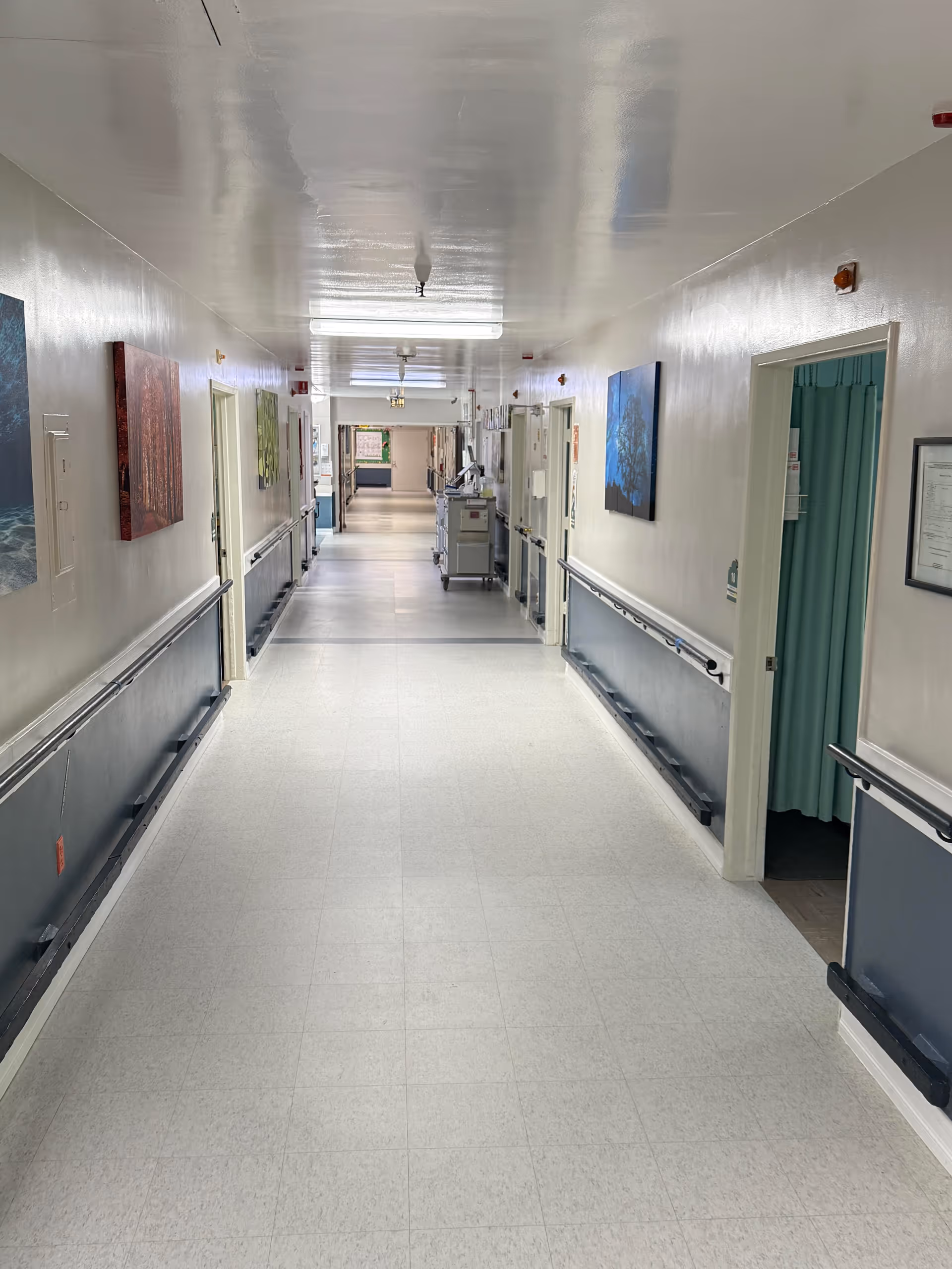 Long, well-lit hospital-style corridor with doors on both sides and wall rails.