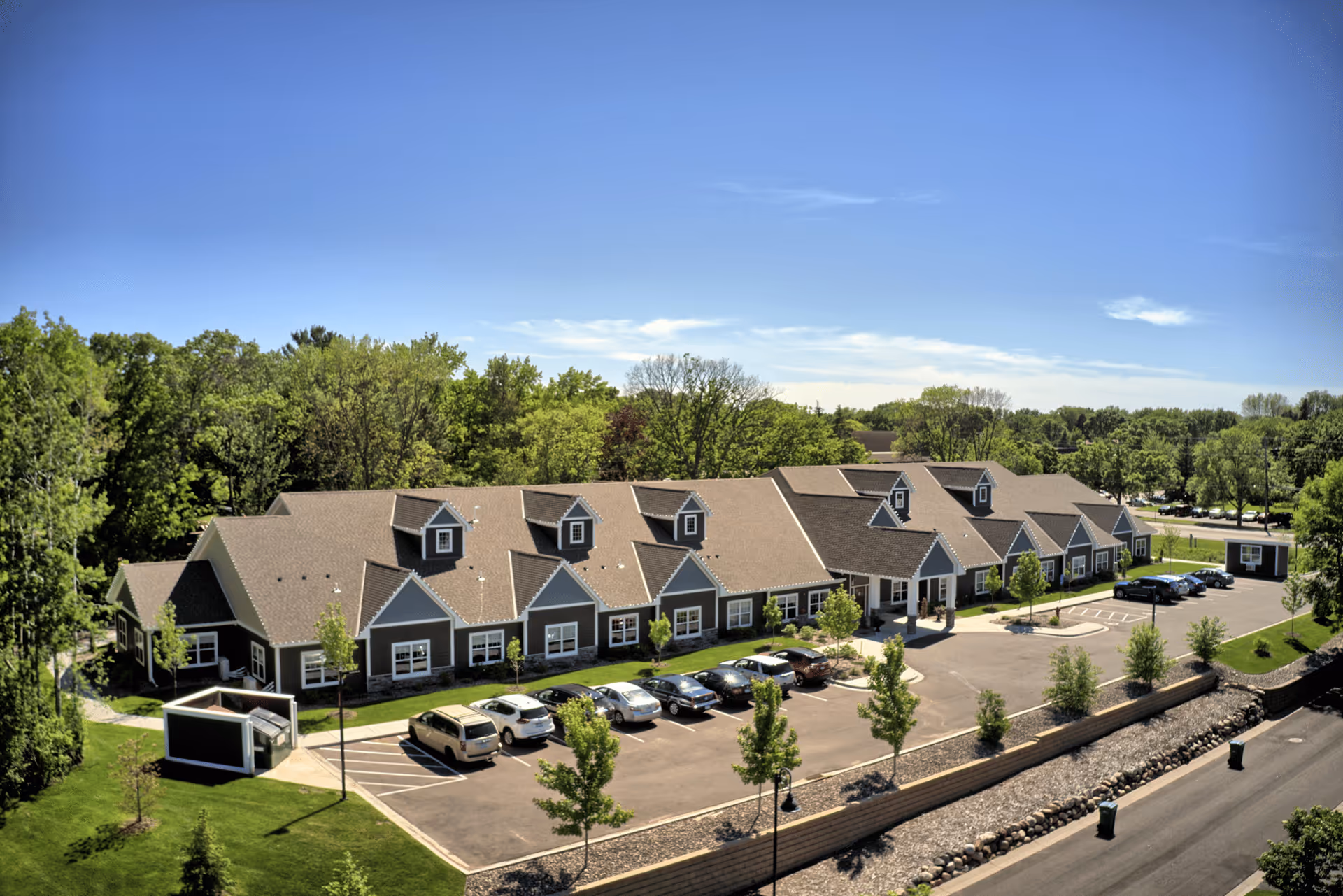 Aerial view of a long single-story senior living building with dormer windows, parked cars, and surrounding trees under a clear blue sky.