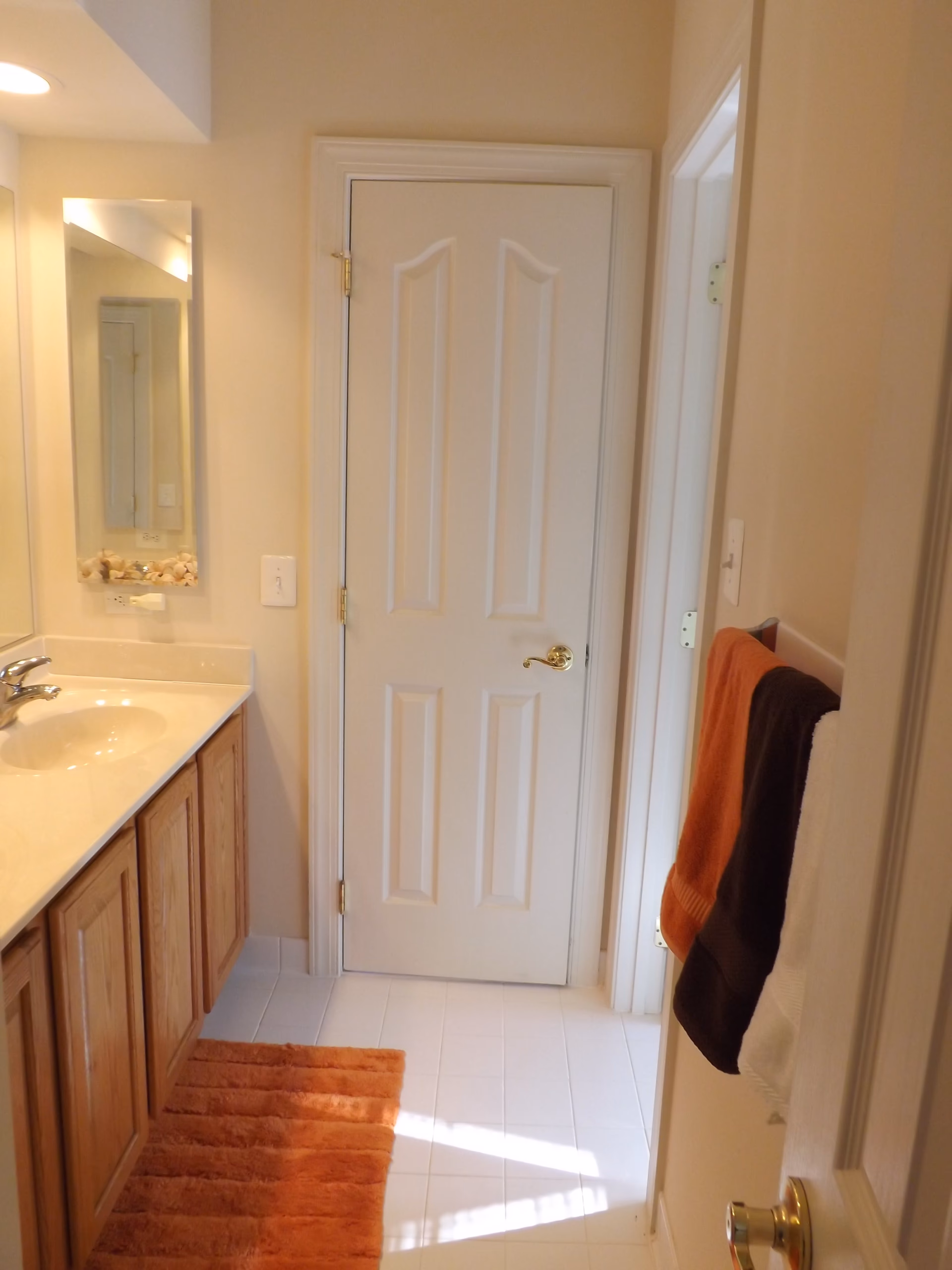 A bathroom interior with a white door at the center, a wooden vanity with a sink and faucet on the left, a rectangular mirror above the sink, and three towels (orange, brown, and white) hanging on a towel rack on the right wall. There is an orange bath mat on the white tiled floor.