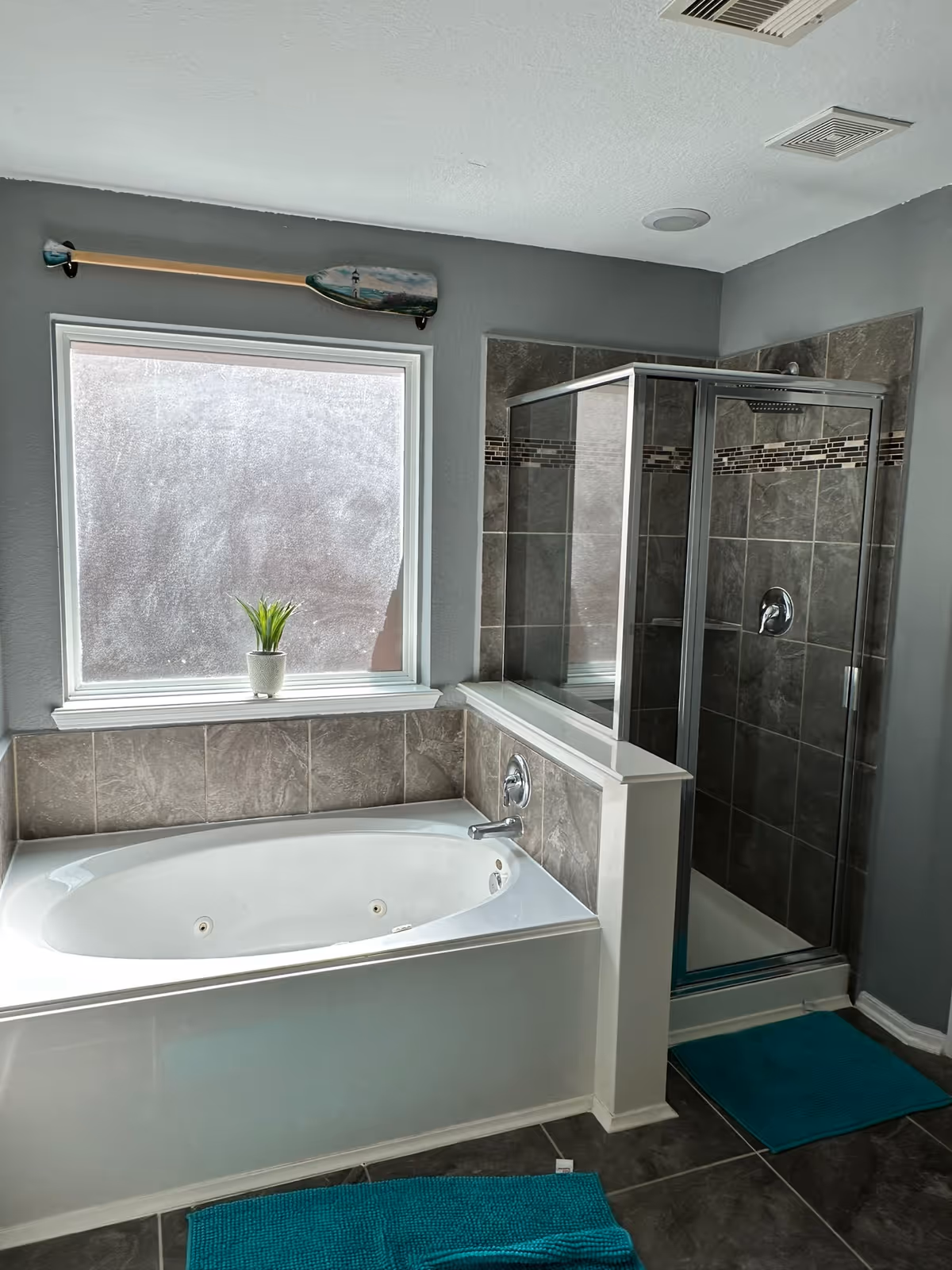 Bathroom with a jetted tub, glass shower, gray tiles, and a plant on the window sill.