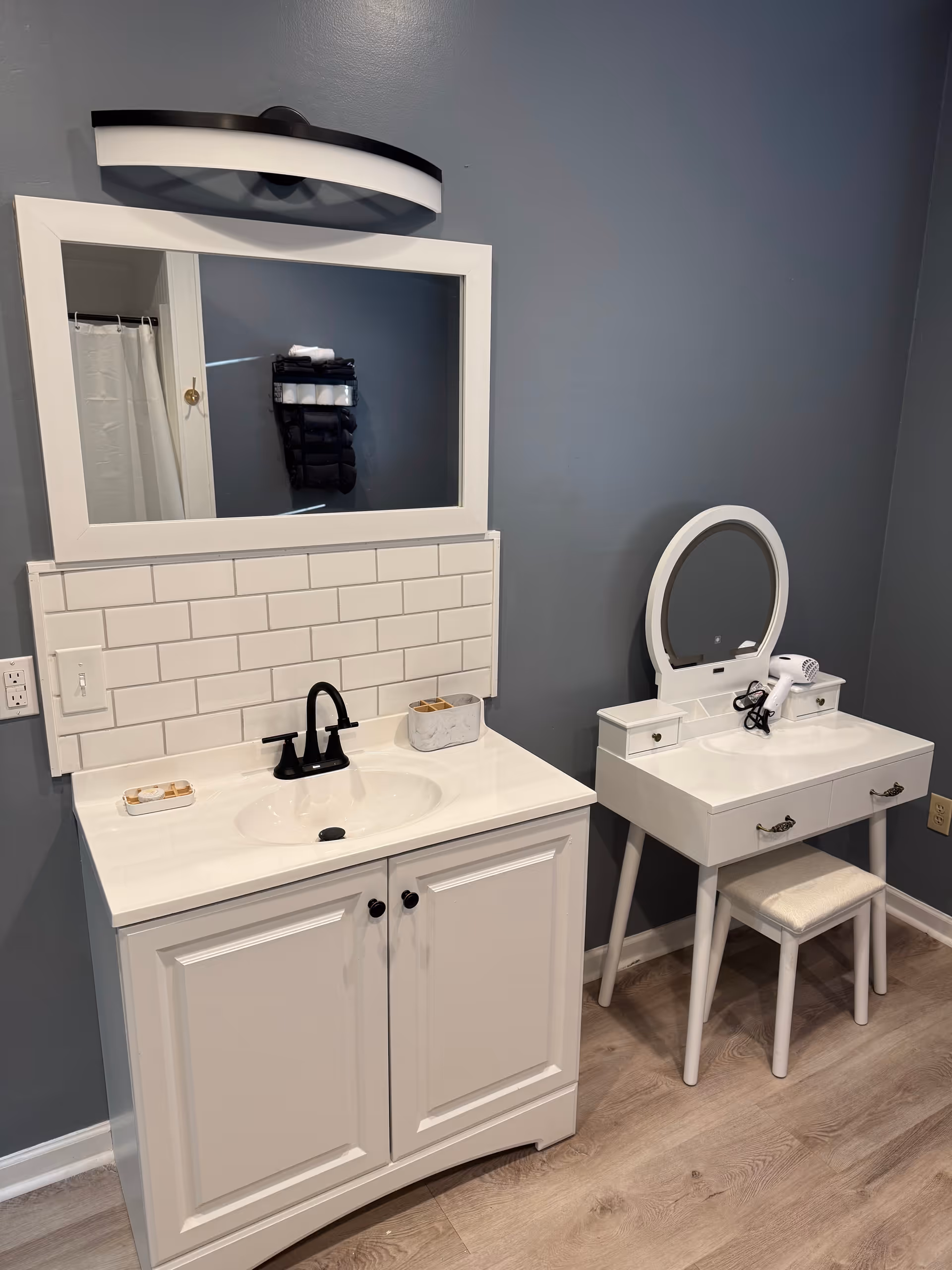 Blue-walled bathroom with white vanity, mirror, tiled backsplash, and a small dressing table.