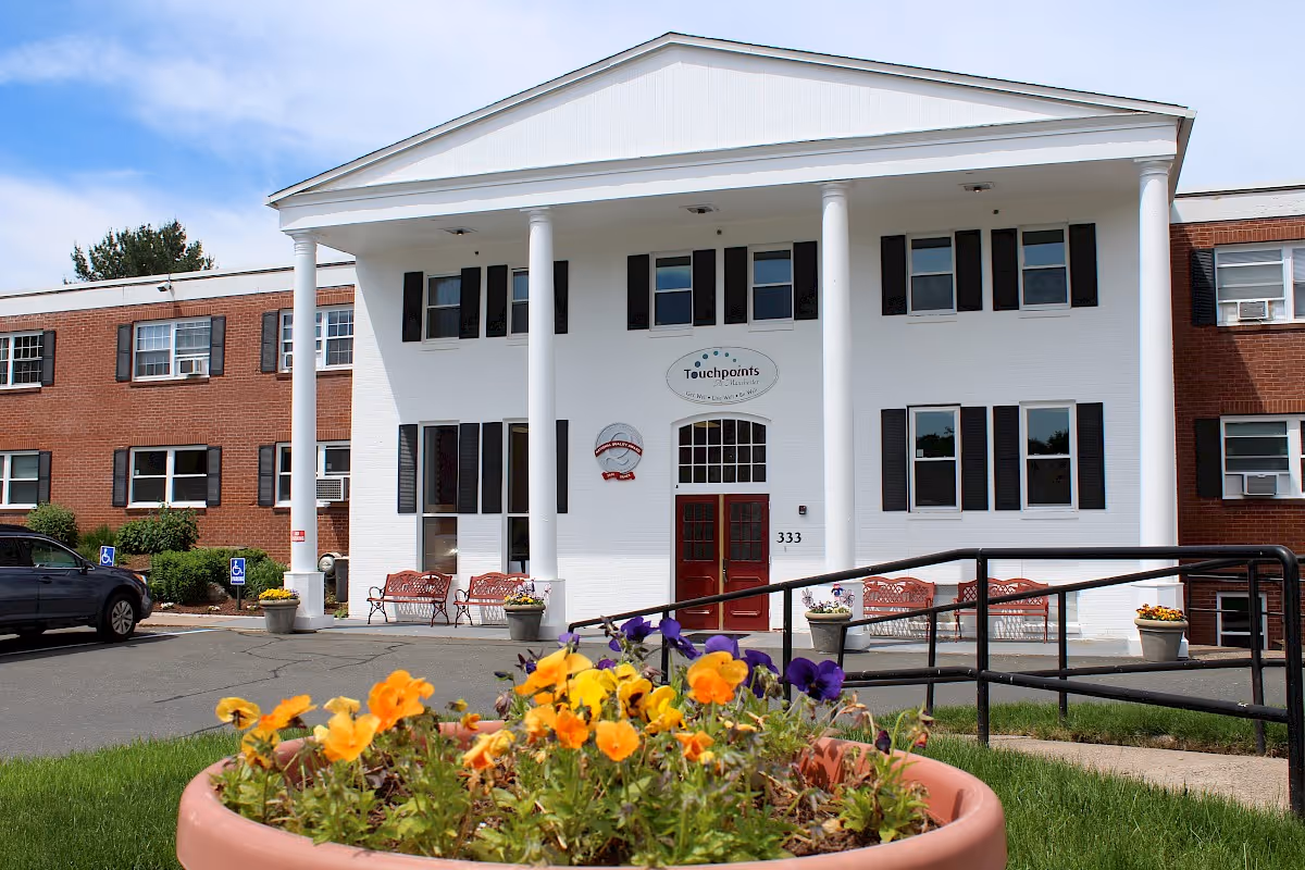 Front entrance of a brick-and-white senior living building with large white columns, red doors, benches, and a flower planter in the foreground.