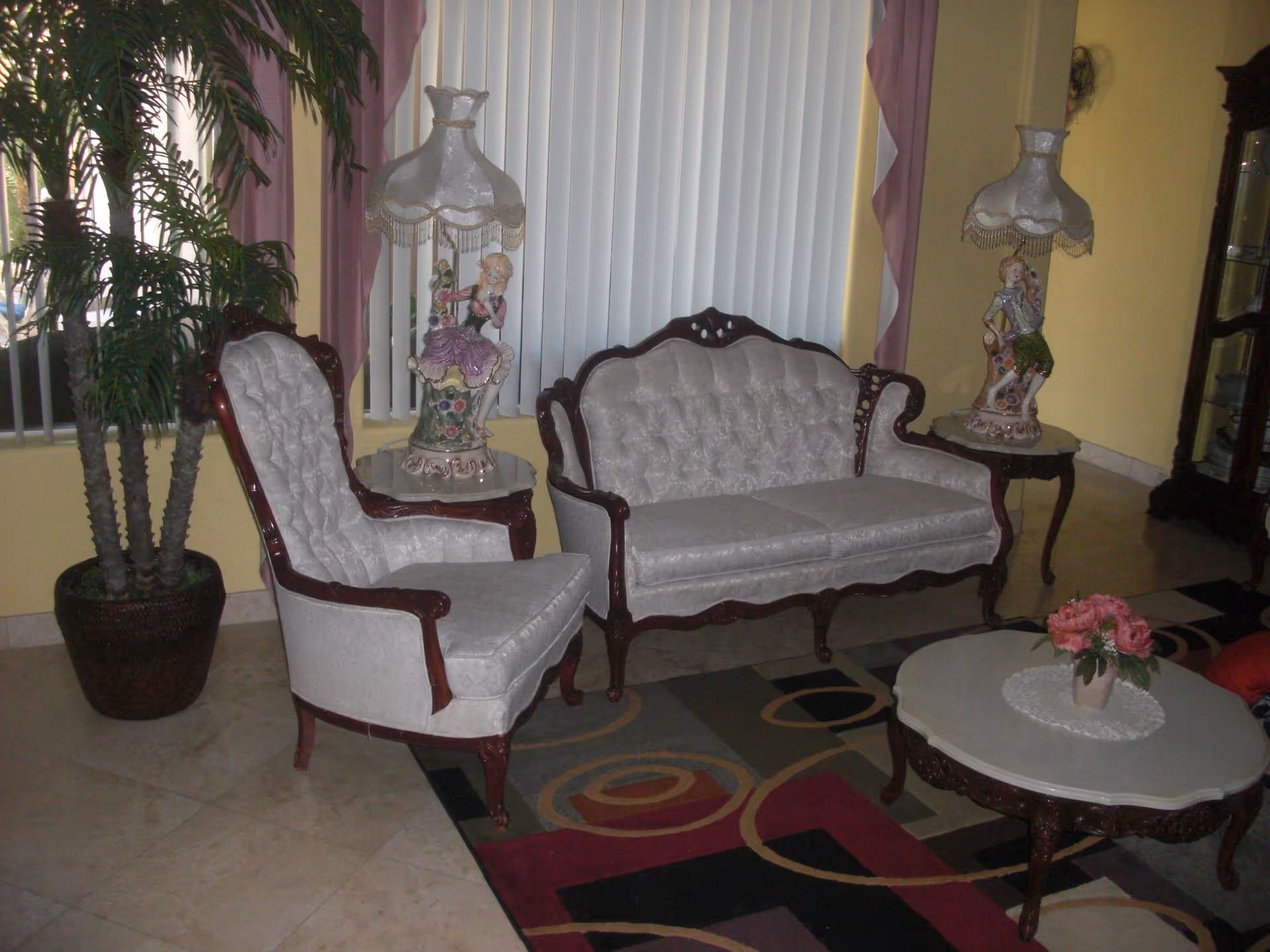 A living room with vintage-style white upholstered wooden sofa and armchair, two decorative table lamps with figurine bases on side tables, a coffee table with a small flower arrangement, a colorful patterned rug, a large potted plant, and vertical blinds covering the window.