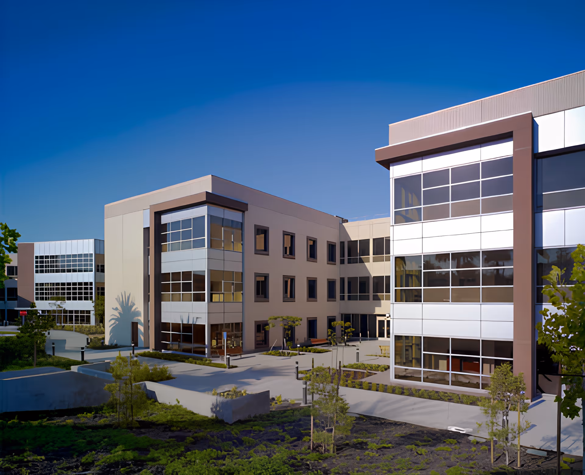 Modern multi-story building with large windows surrounding a landscaped courtyard under a clear blue sky.
