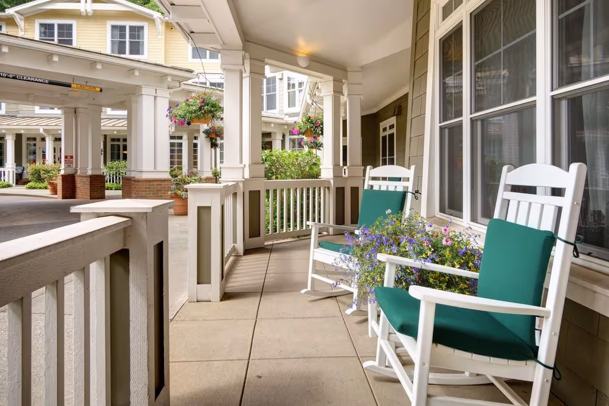 Covered front porch of a senior living facility with white rocking chairs with green cushions, potted and hanging flowers, and a driveway canopy.