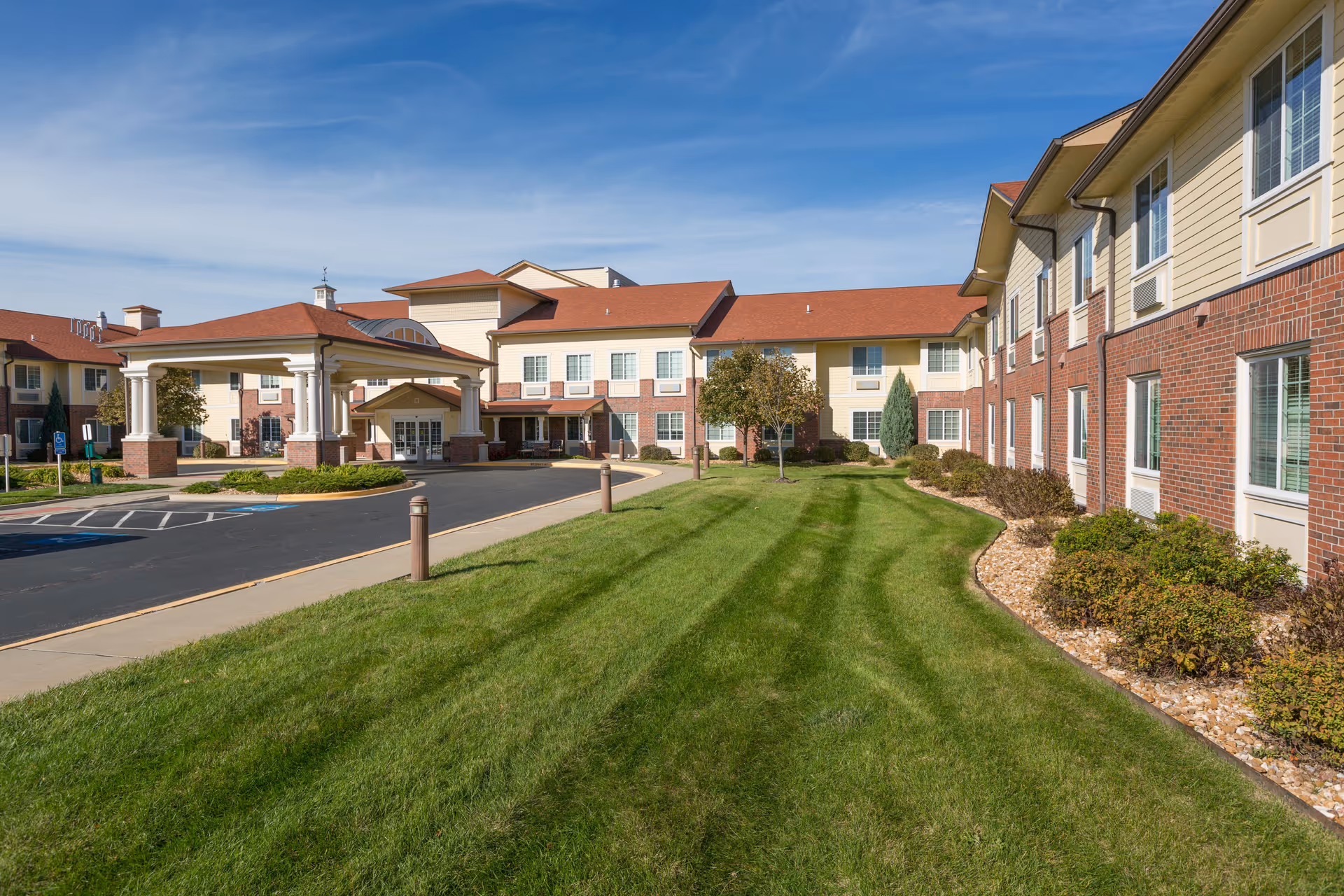 Exterior view of a senior living facility with a well-maintained lawn, shrubs, and a paved driveway leading to a covered entrance. The building has a combination of brick and light-colored siding with multiple windows and a red roof under a clear blue sky.